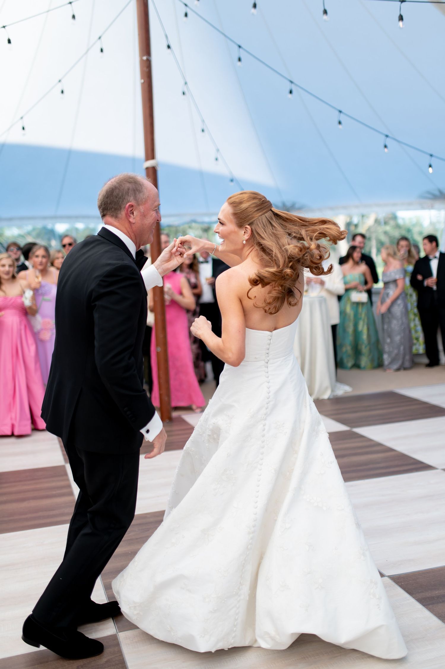 Bride in strapless gown dancing with father under string lights at outdoor tented reception.