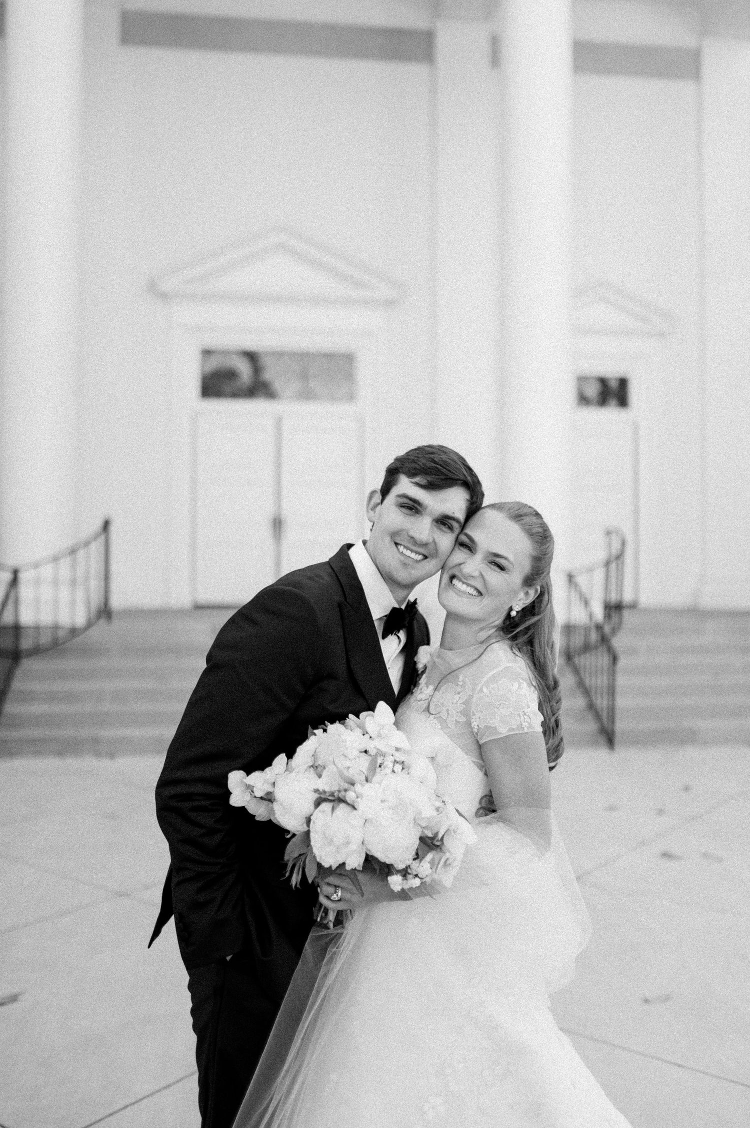 Black and white portrait of smiling bride and groom embracing outside a classic white columned church.