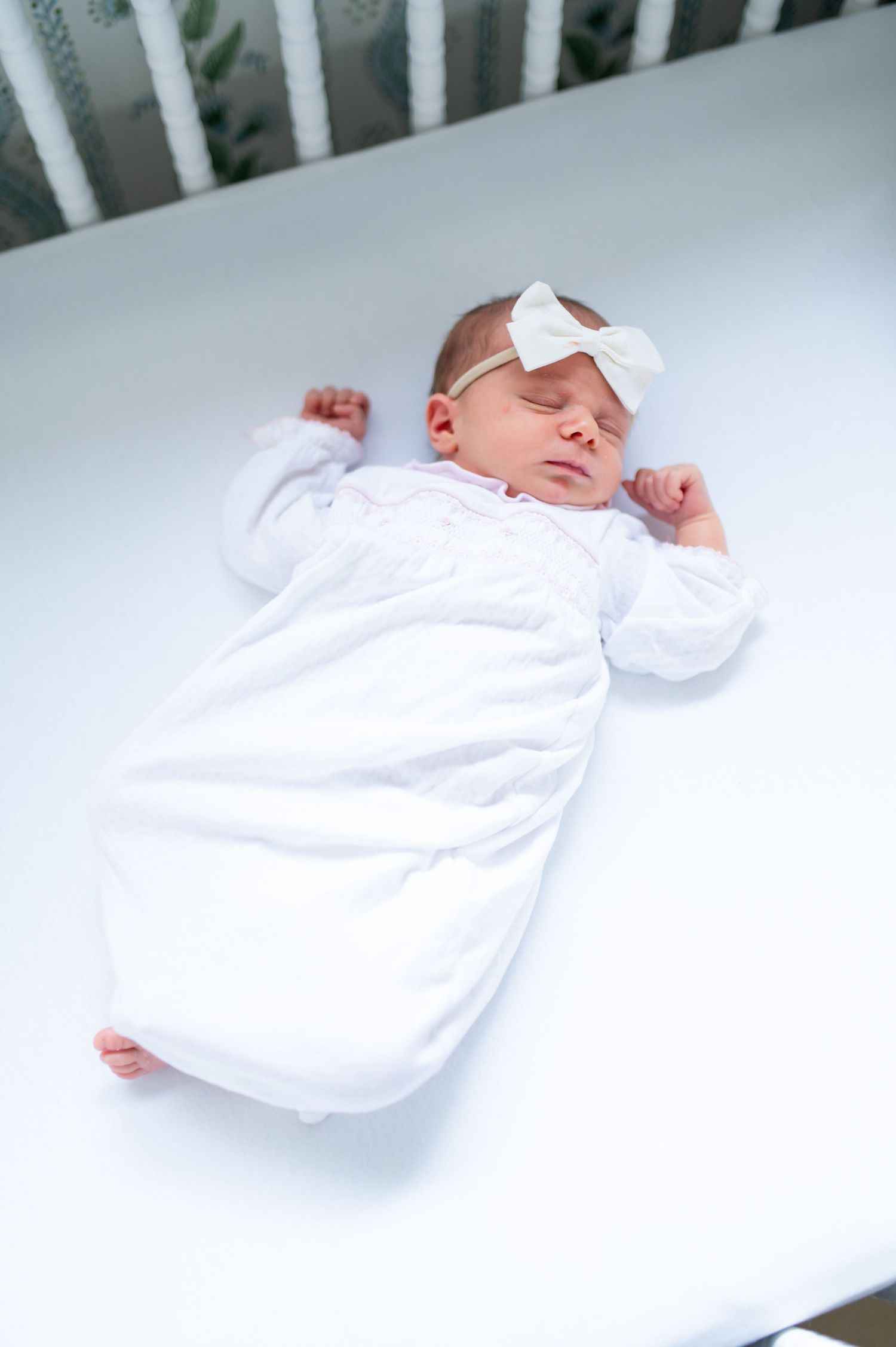 A sleeping newborn baby wearing a white outfit and bow headband rests peacefully in a white crib.