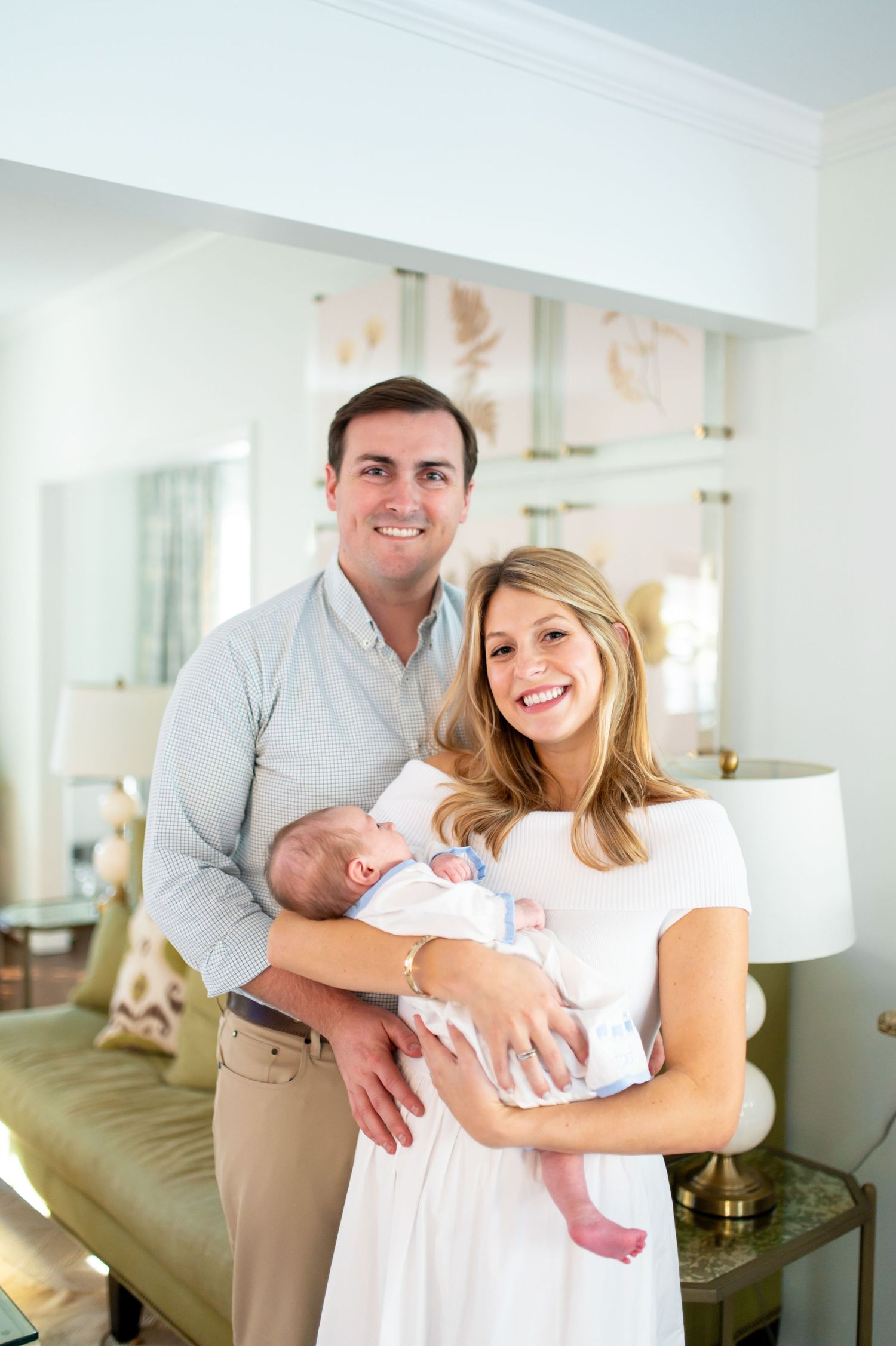 A couple stands in a bright living room holding their newborn baby while smiling at the camera.