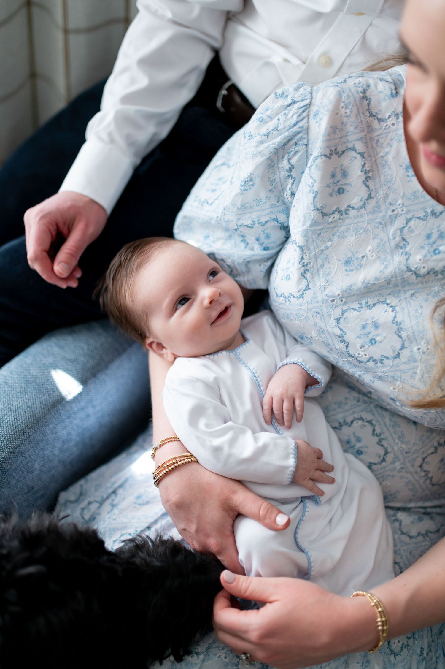 A baby in a white outfit rests on someone's lap wearing denim jeans while being held with gentle hands.