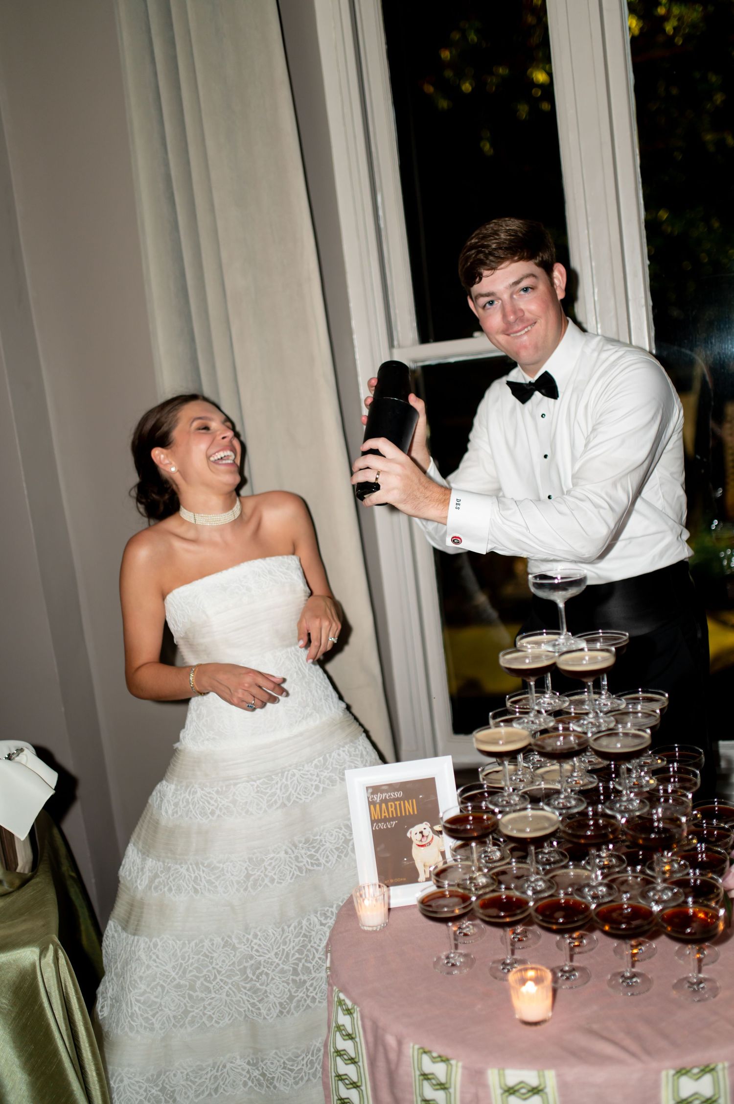 Groom pours espresso martini tower as bride laughs joyfully at elegant wedding reception.