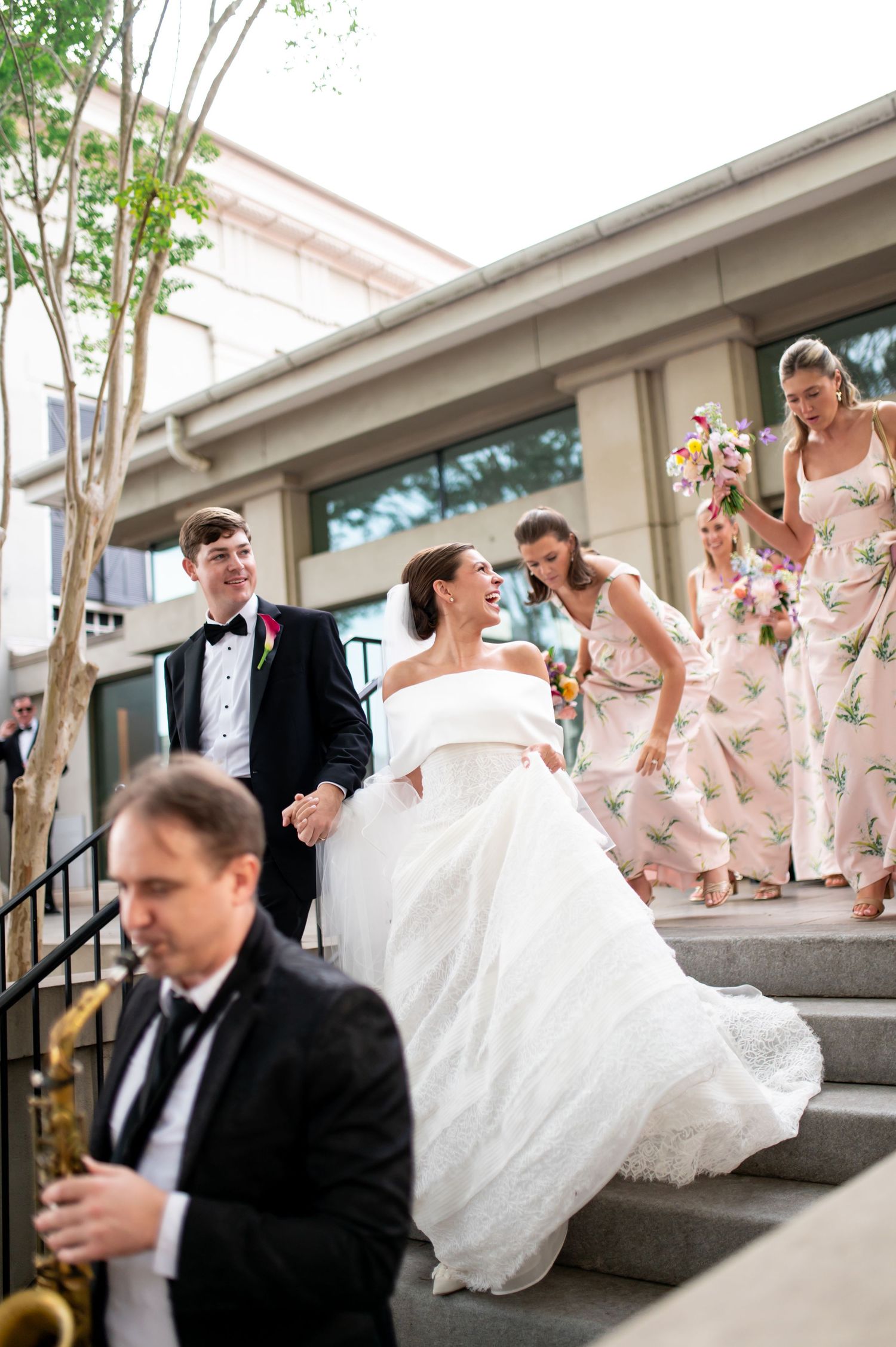 Bride and groom descend steps with bridesmaids in floral dresses as saxophonist plays at wedding.