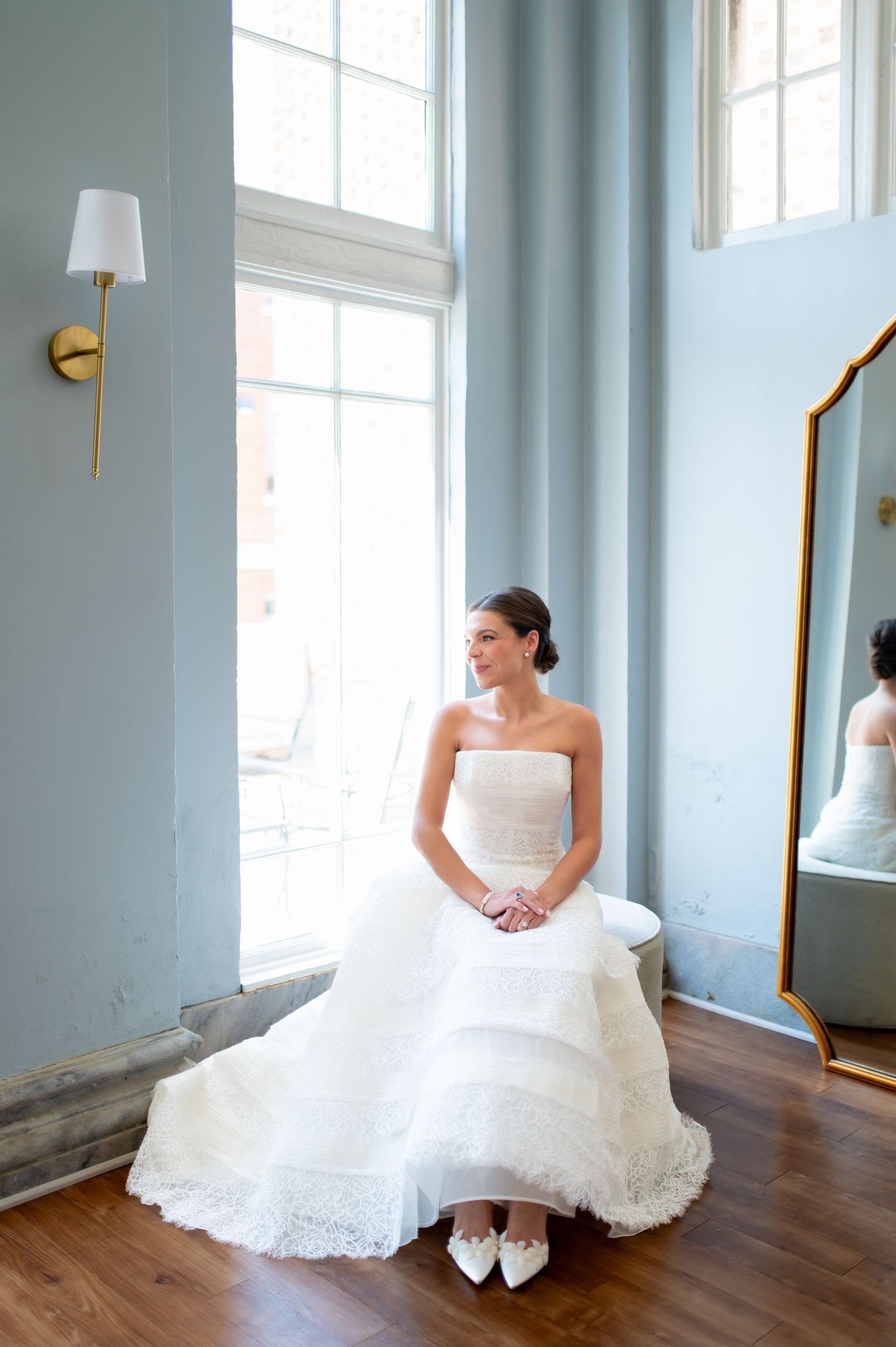 Elegant bride in strapless white lace ball gown sits near tall windows beside a gold ornate floor mirror.