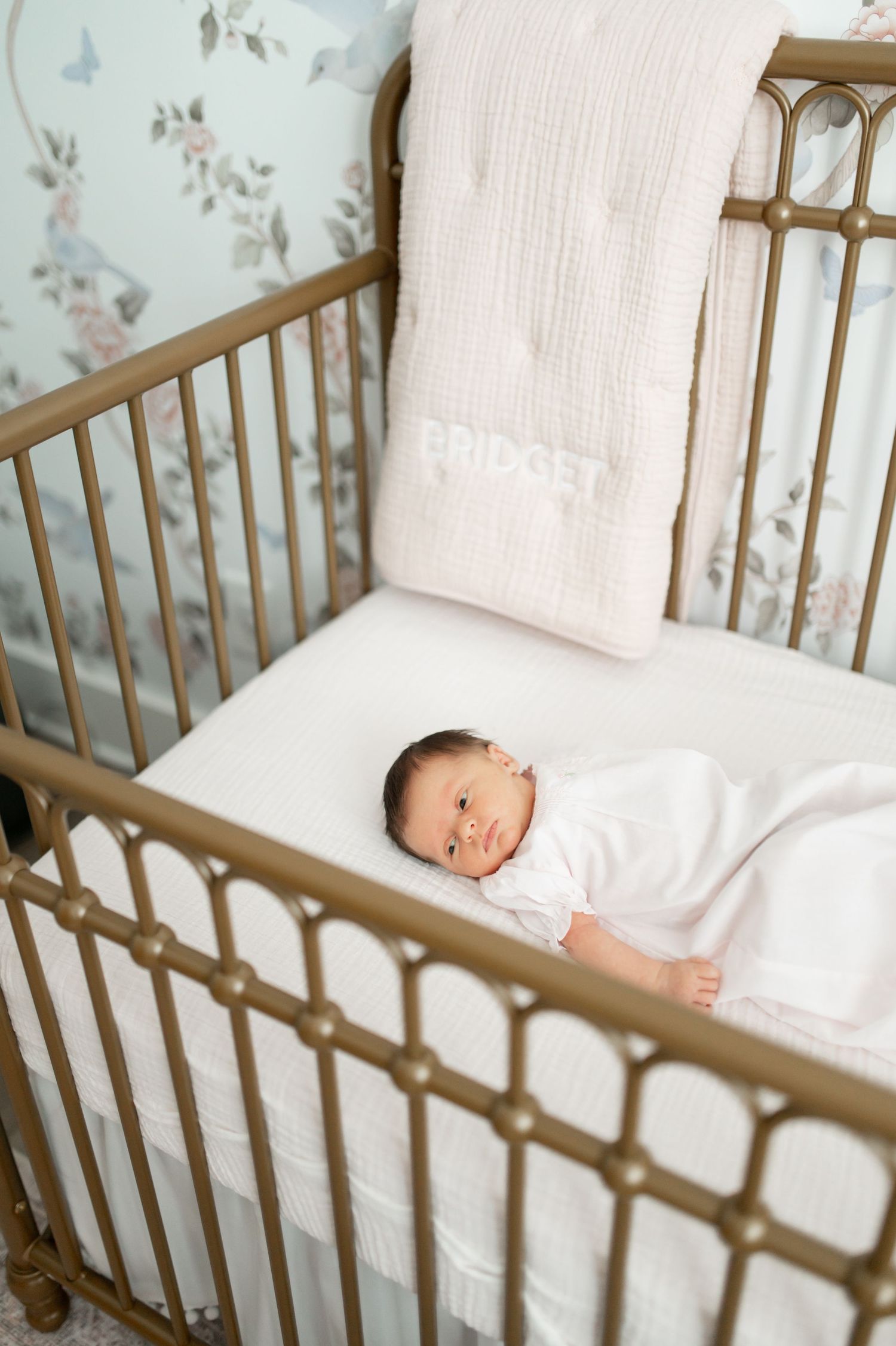 Antique brass baby crib with white bedding against soft floral wallpaper.