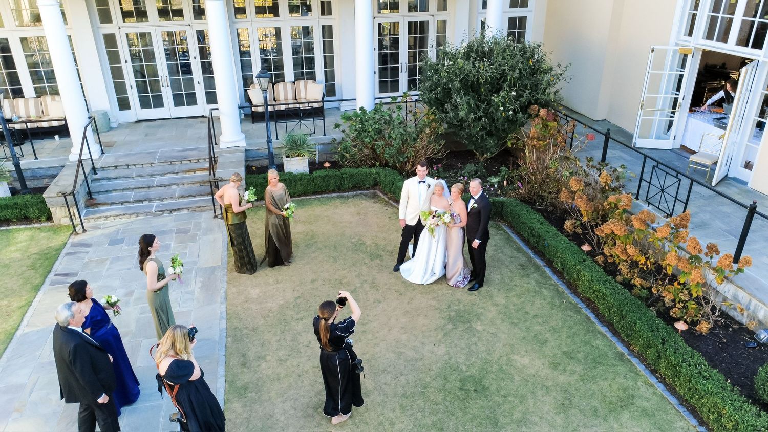 Aerial view of a wedding party posing on a green lawn outside an elegant venue with bridesmaids in gold gowns.