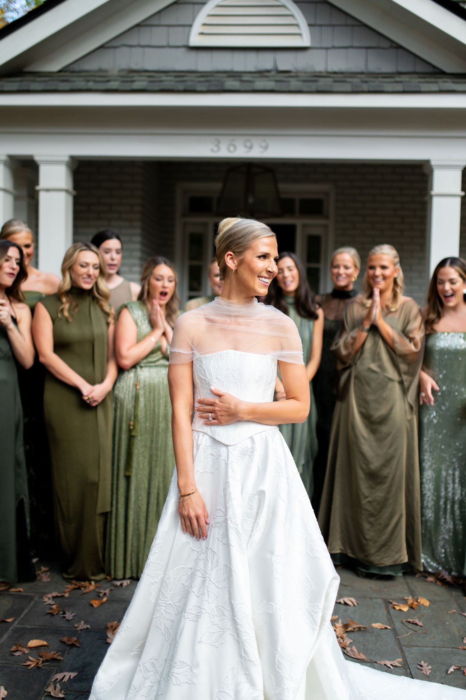 Wedding party in sage green dresses poses with someone in a white gown on a front porch.