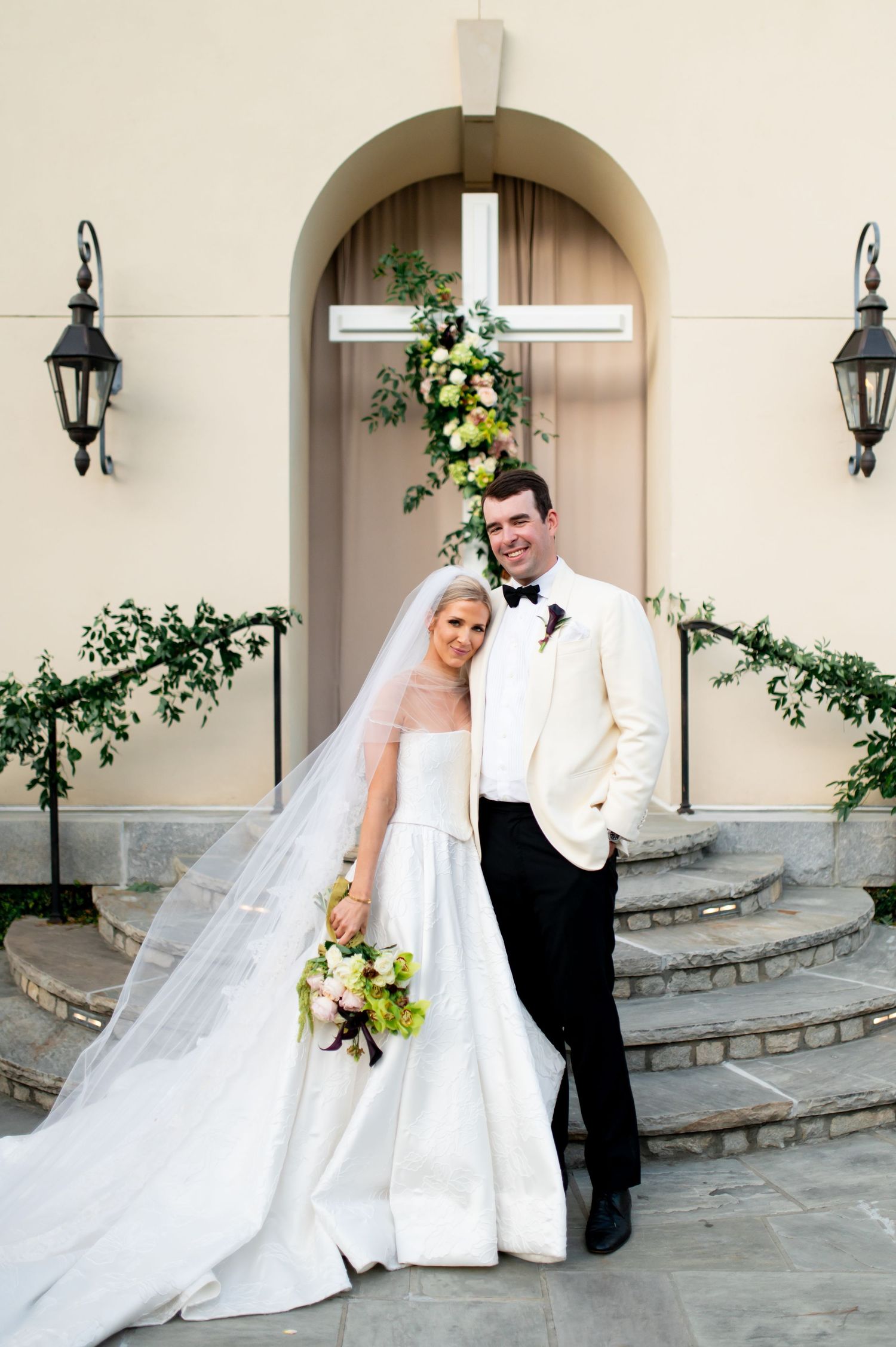 Bride in flowing white gown and veil poses with groom in white dinner jacket.