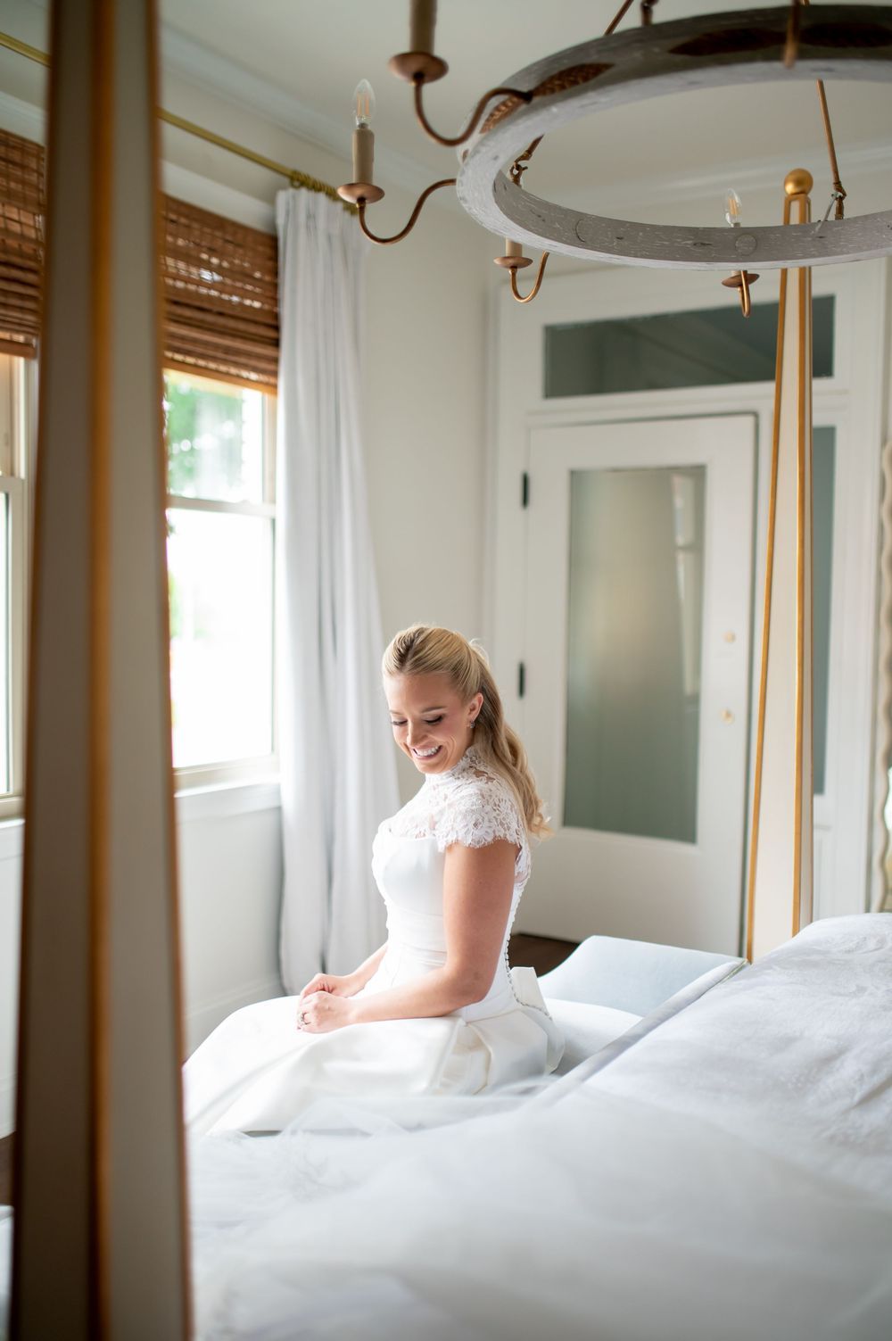Smiling bride in a white lace wedding dress sitting on a bed in an elegant room.