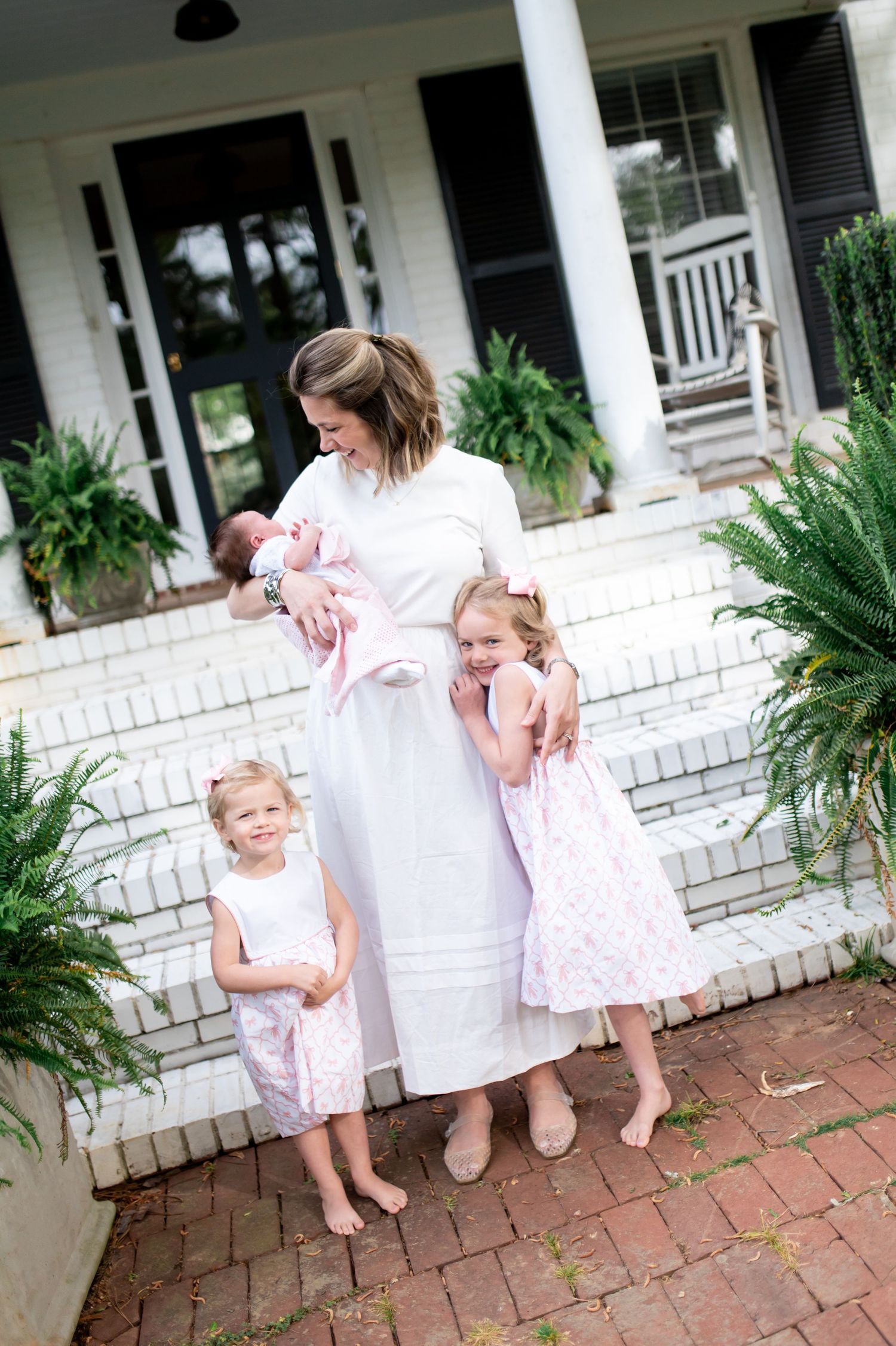 A mother in a white dress holds a newborn while two young girls in pink dresses hug her on brick steps.