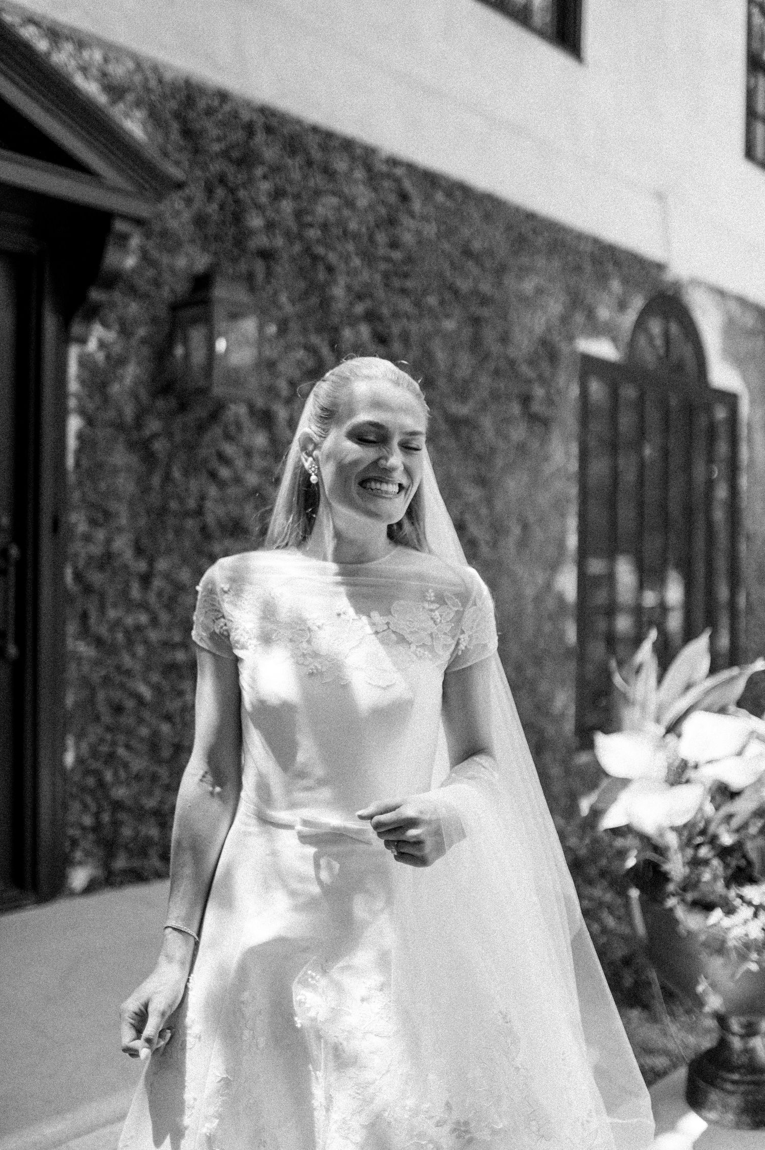 Joyful bride in lace wedding gown and long veil laughing outdoors by ivy-covered wall.