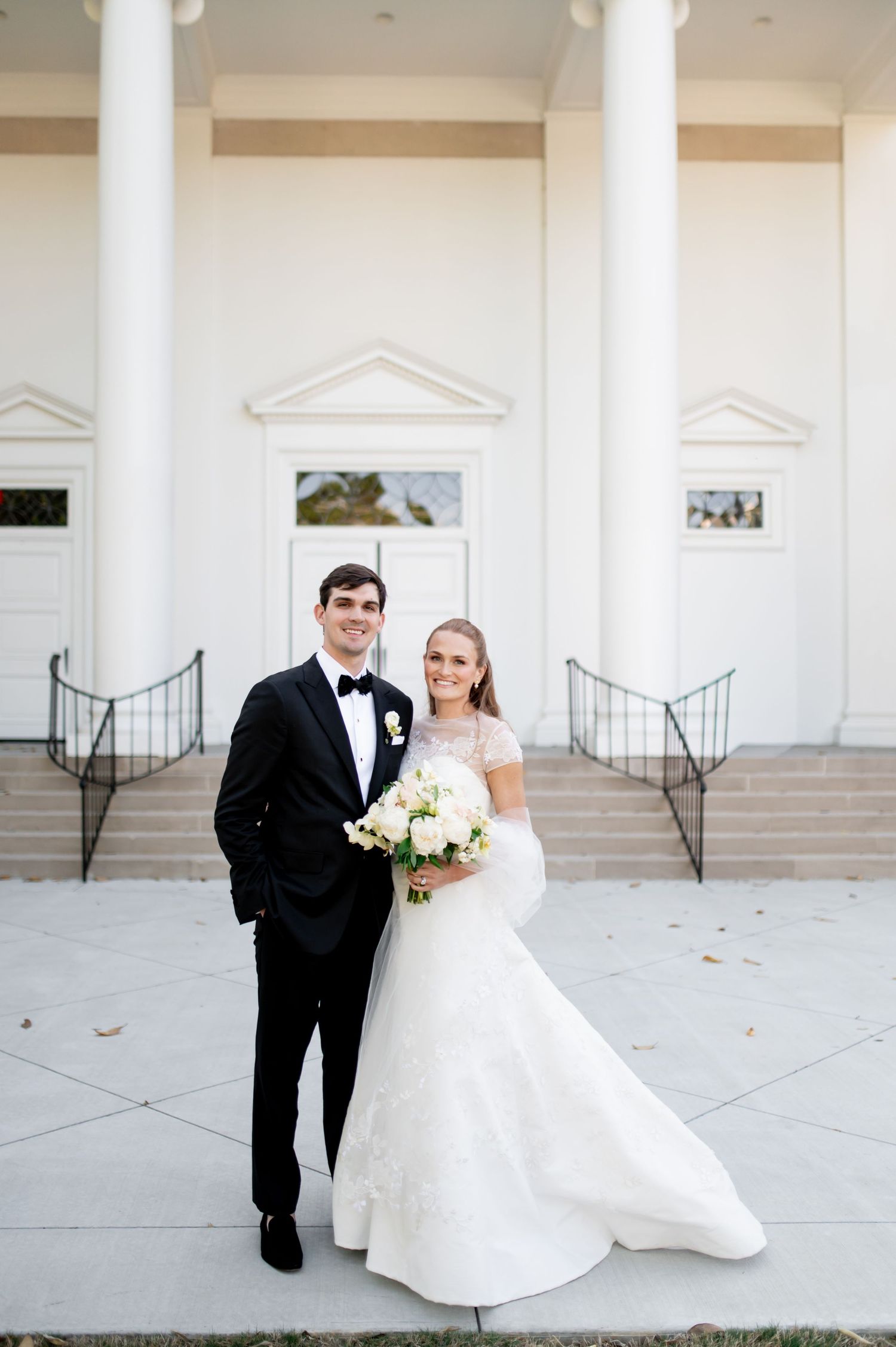Bride and groom pose before white columned church entrance, bride holding white bouquet in fitted gown.
