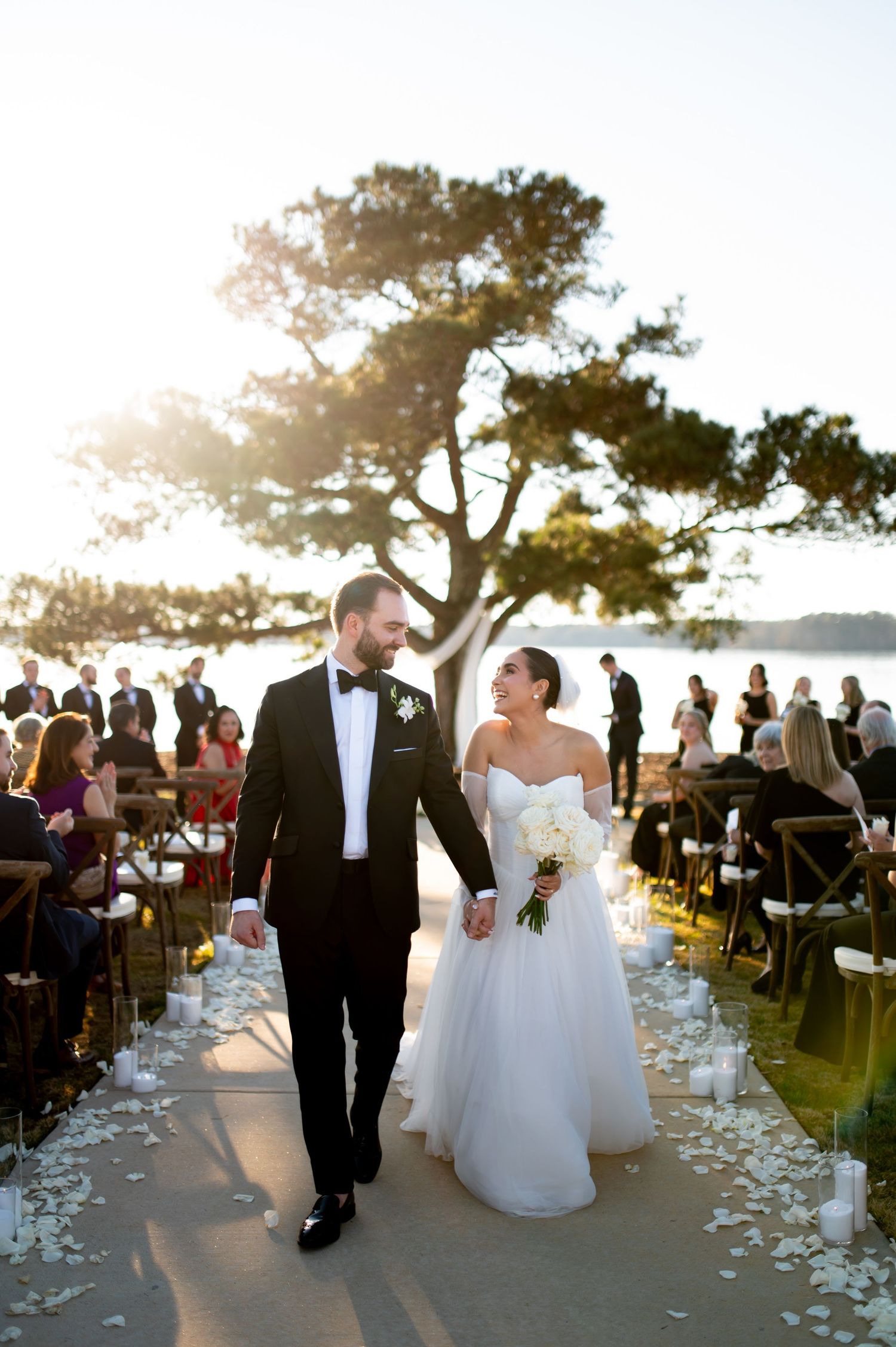 Bride and groom walk down outdoor aisle at sunset waterfront wedding ceremony, guests seated beside rose petal lined path.