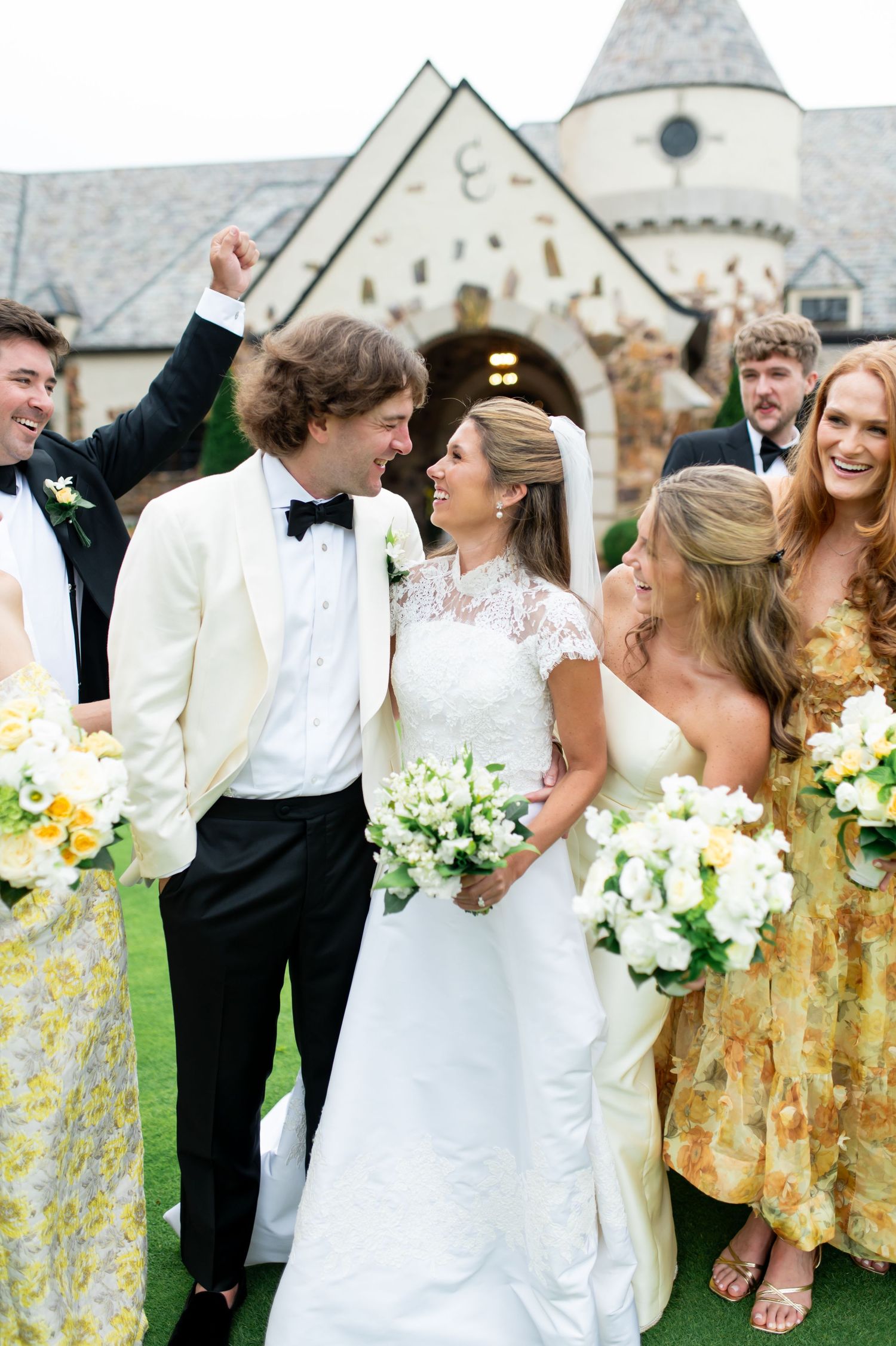 Wedding party in yellow floral dresses and formal wear celebrate on lawn near stone building.