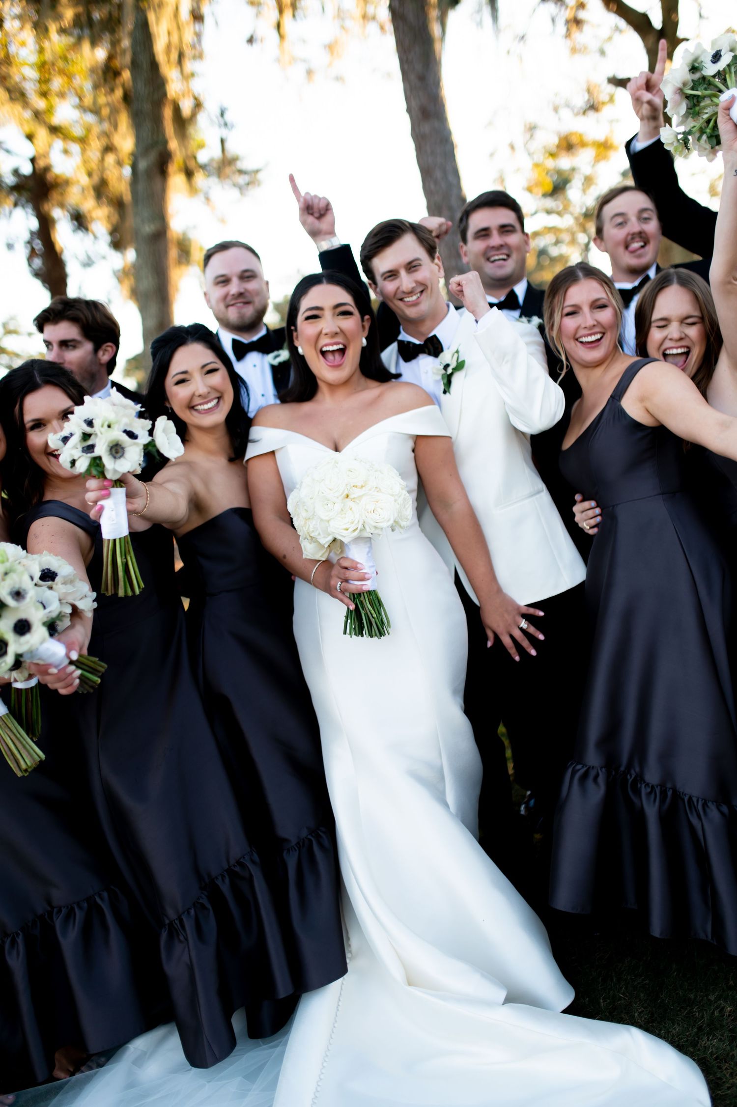 Bridal party in black dresses and white gown poses energetically outdoors under trees.