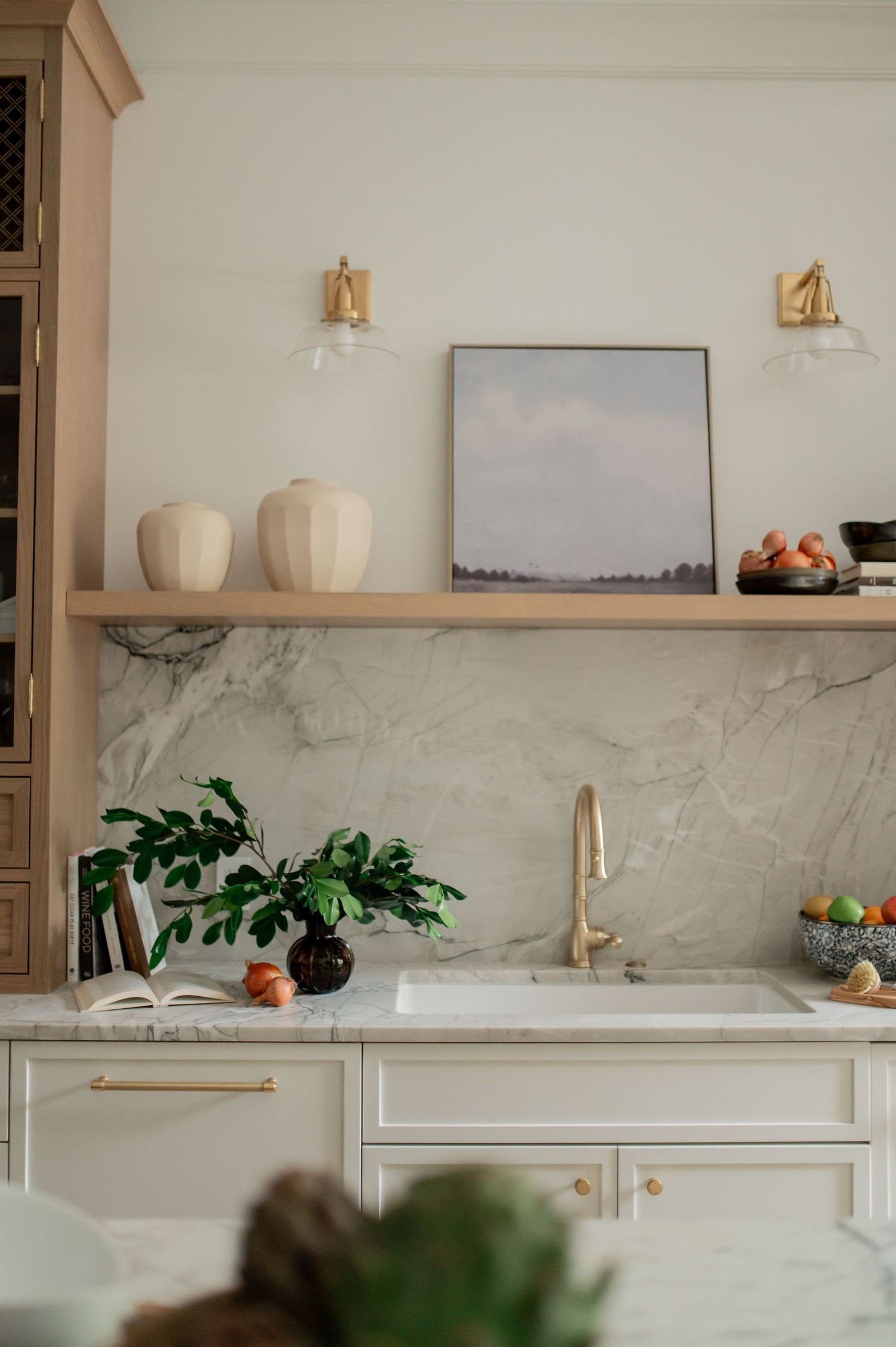 Elegant kitchen with marble backsplash, brass faucet, open wood shelf with vases and art, and white cabinetry with gold hardware.