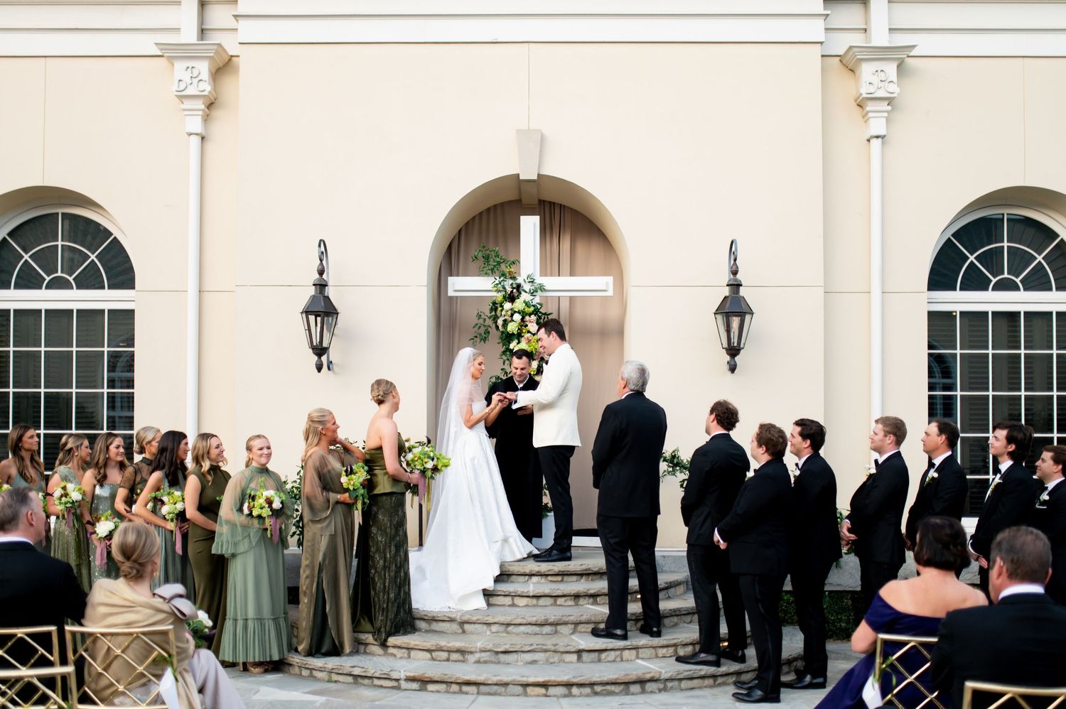 Wedding ceremony setup with guests seated in front of a cream-colored building with arched doorways.