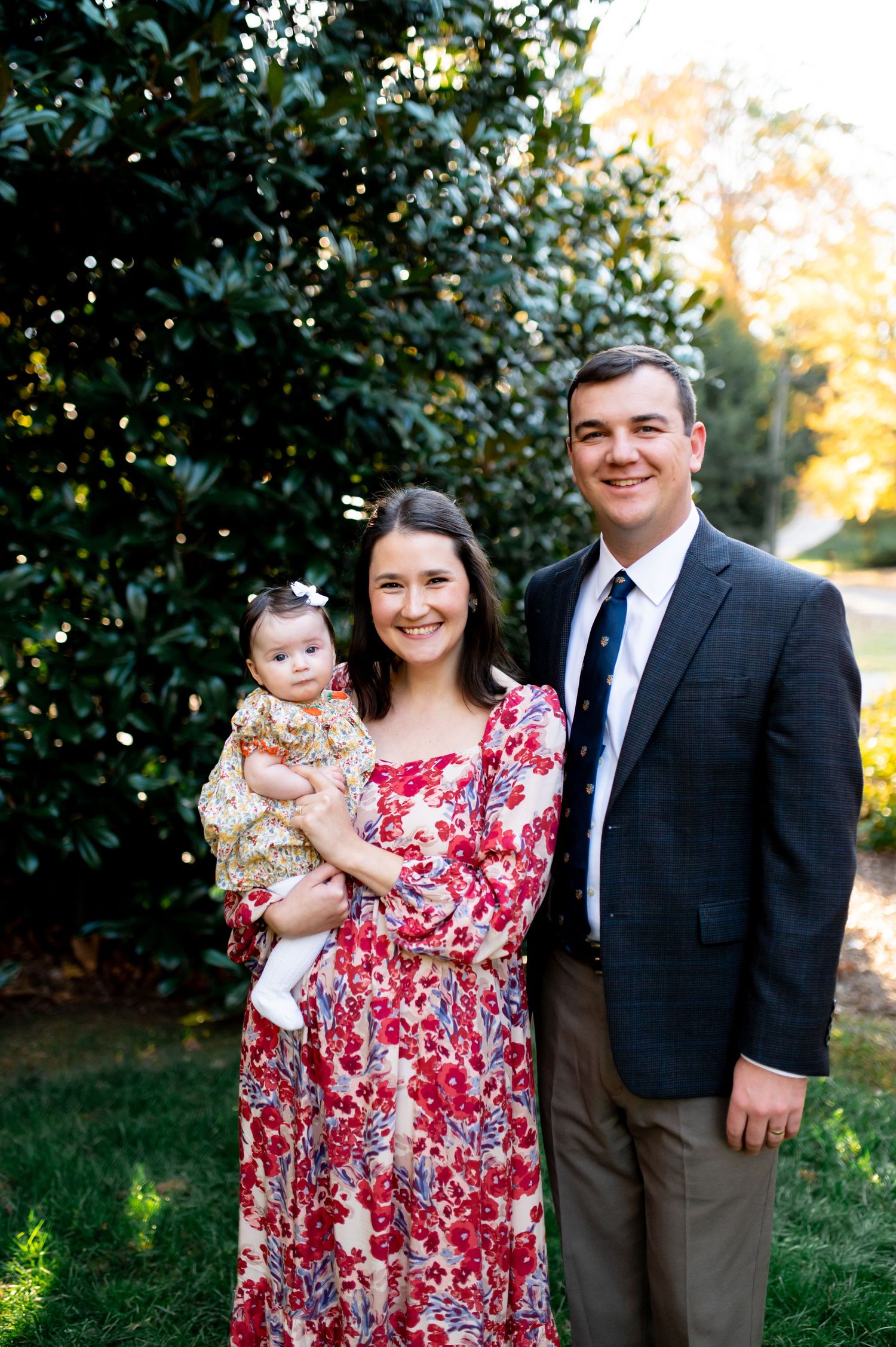 A young family poses outdoors, mother in floral dress holding baby girl, father in suit jacket, green trees behind.