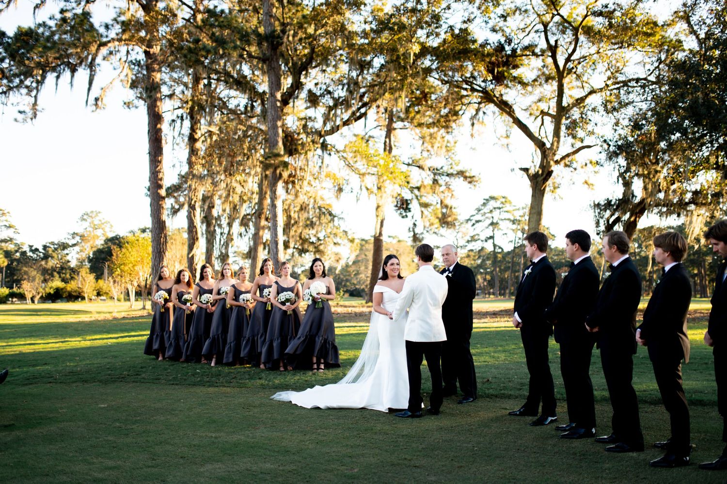 Outdoor wedding ceremony in progress with guests seated in rows under towering oak trees.