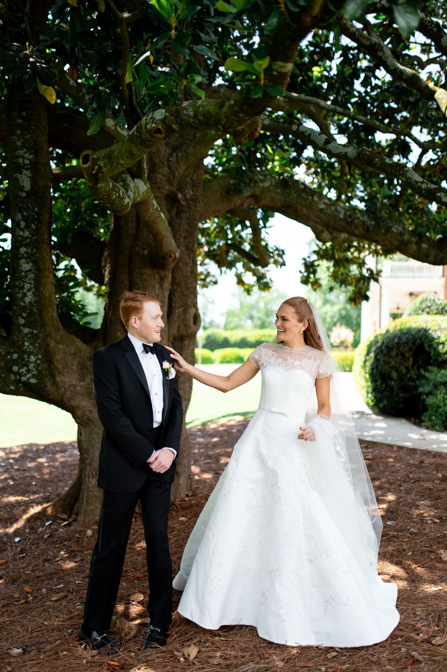 Bride in lace wedding gown touches grooms tuxedo lapel during first look under large oak tree.