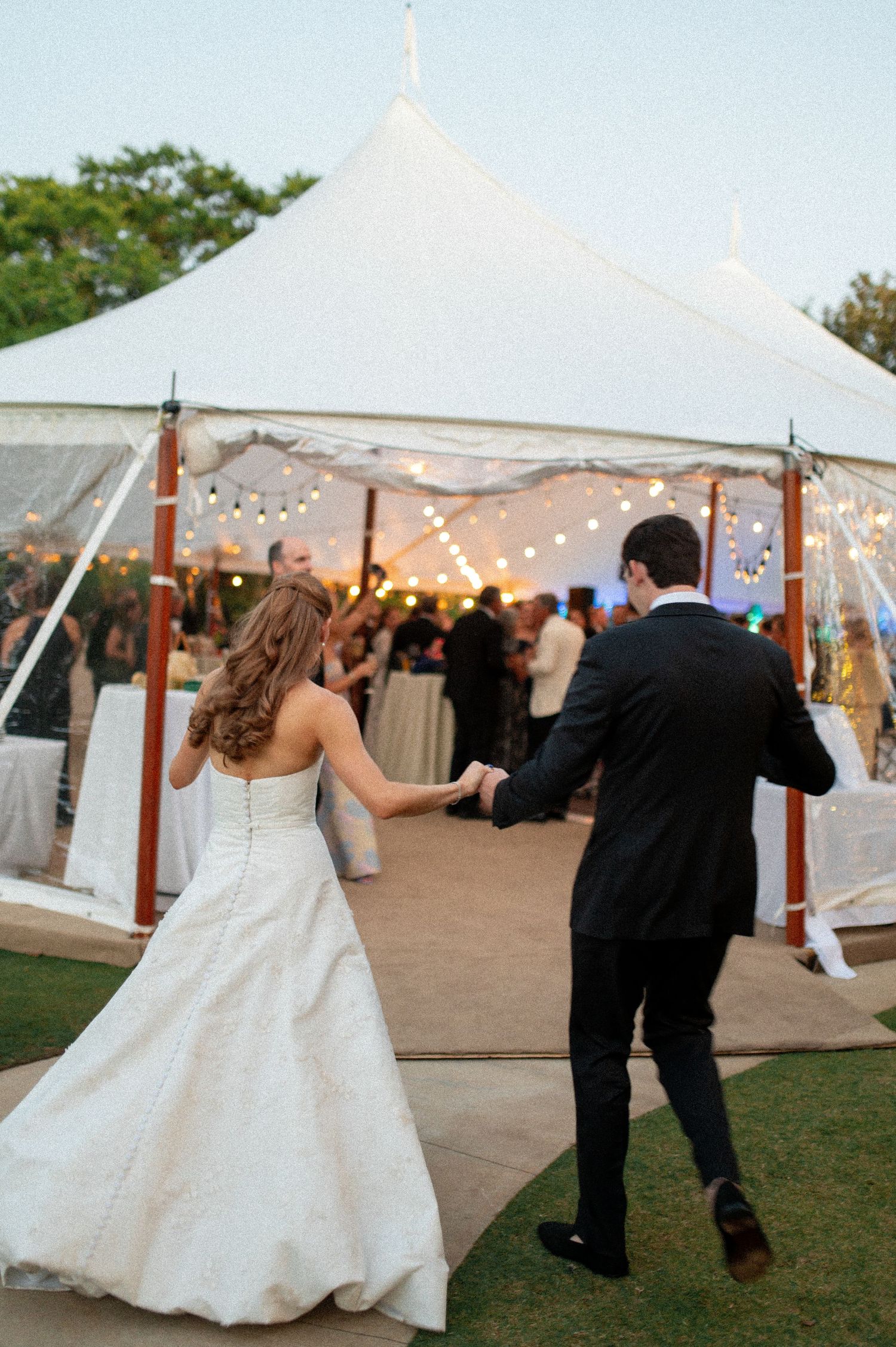 Bride and groom walking hand in hand toward a beautifully lit wedding reception tent at dusk.
