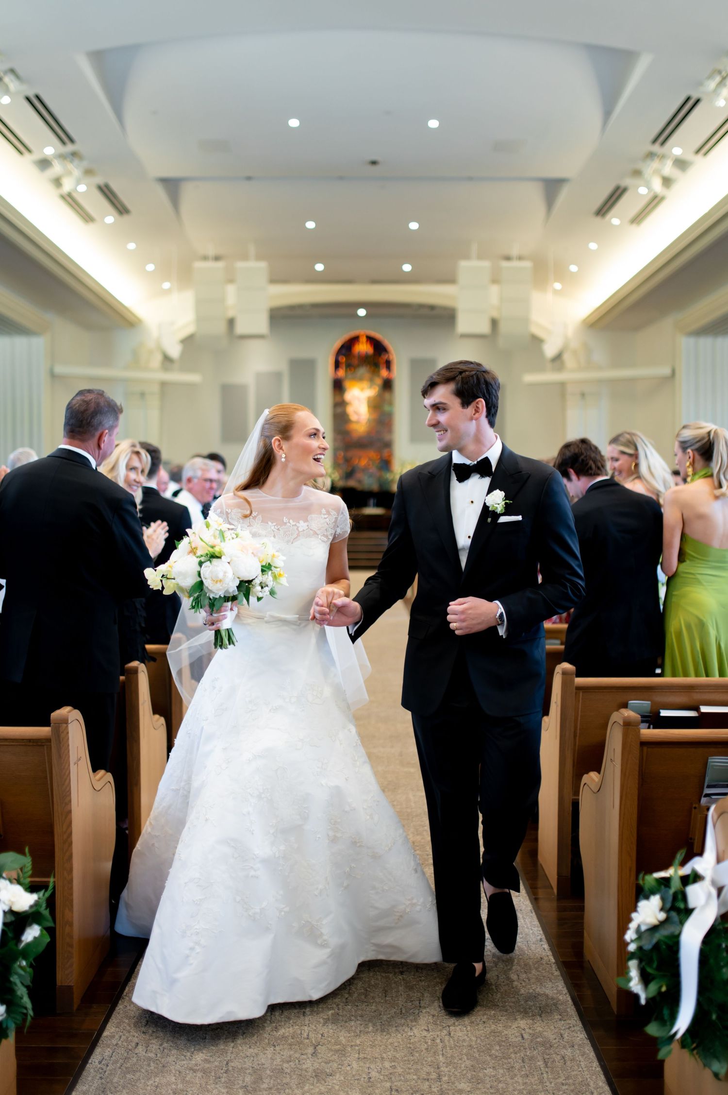 Bride and groom walk down church aisle after wedding ceremony, bride holding white floral bouquet.