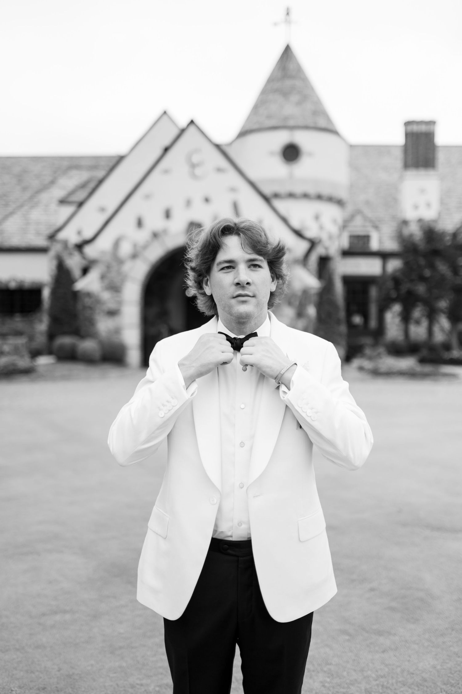 Groom adjusts bowtie while standing in white dinner jacket outside stone church.