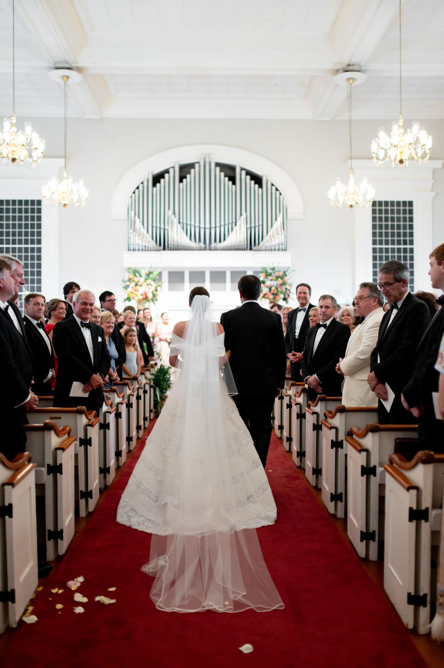 Bride and groom walking down red carpet aisle of elegant white church during traditional wedding ceremony with guests standing.