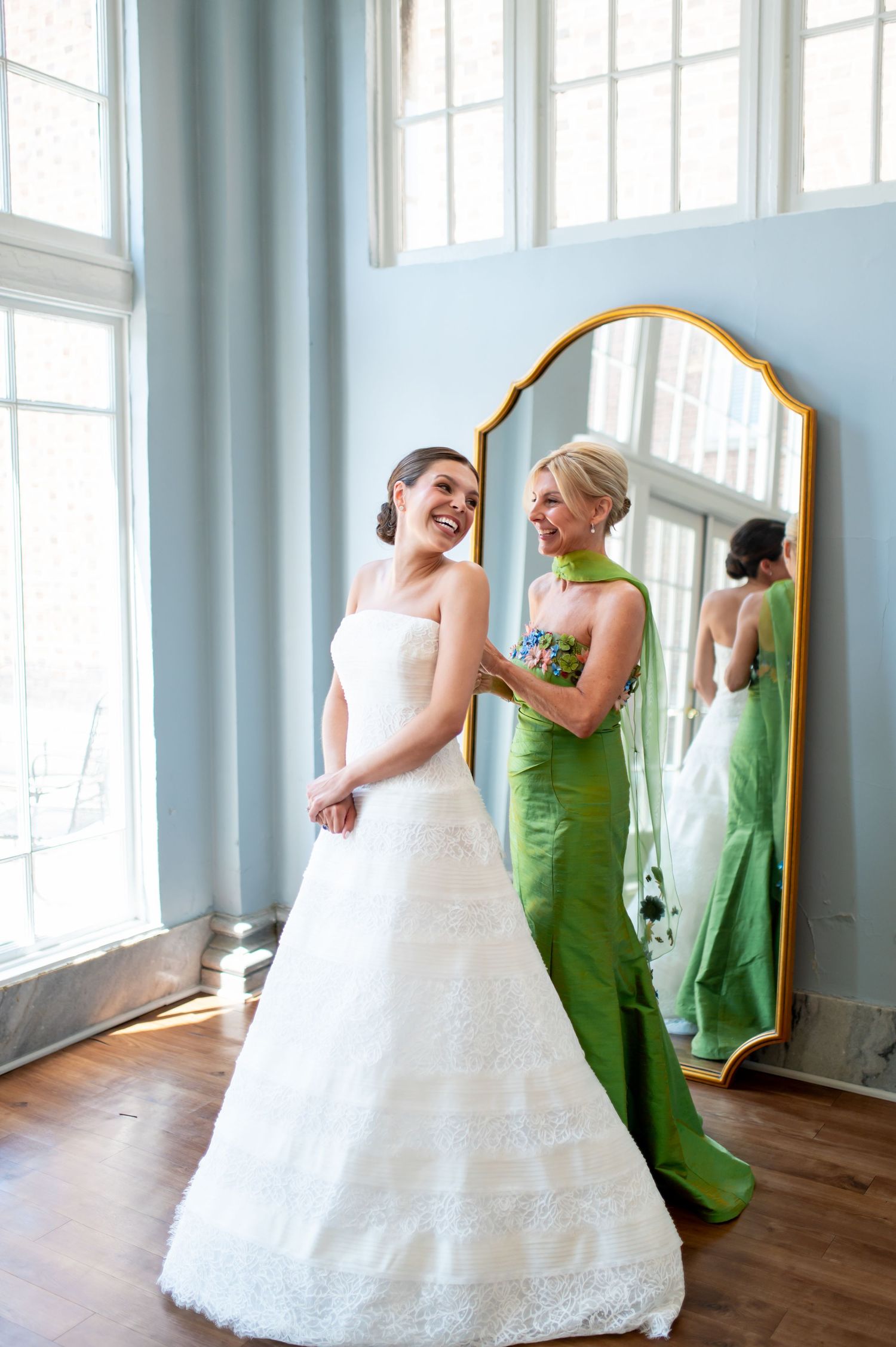 Bride in white strapless gown laughing with bridesmaid in green dress near gold mirror in bright room.