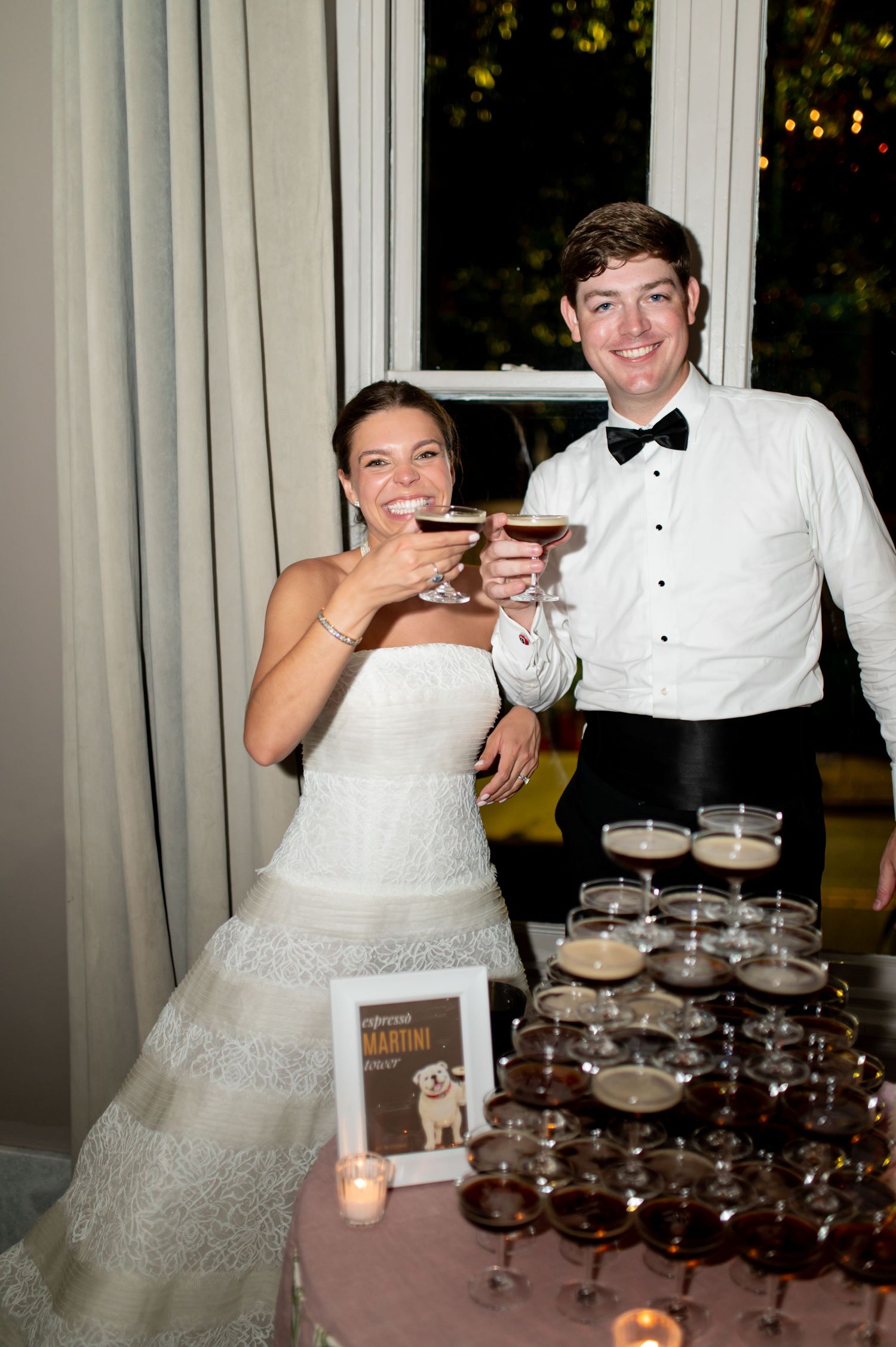 Bride and groom toast with espresso martinis beside a pyramid of cocktail glasses at their wedding.
