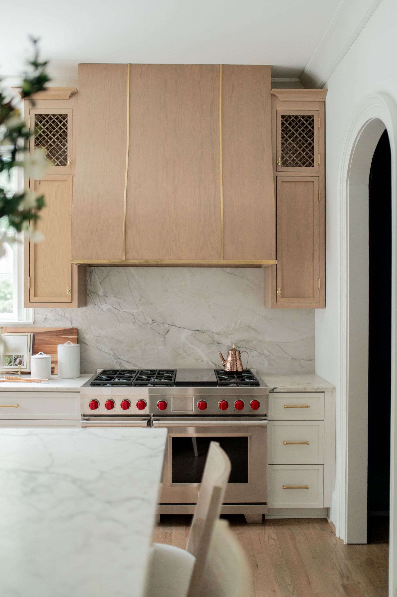 Modern kitchen with light wood range hood, marble backsplash, stainless steel range with red knobs, and gold hardware accents.