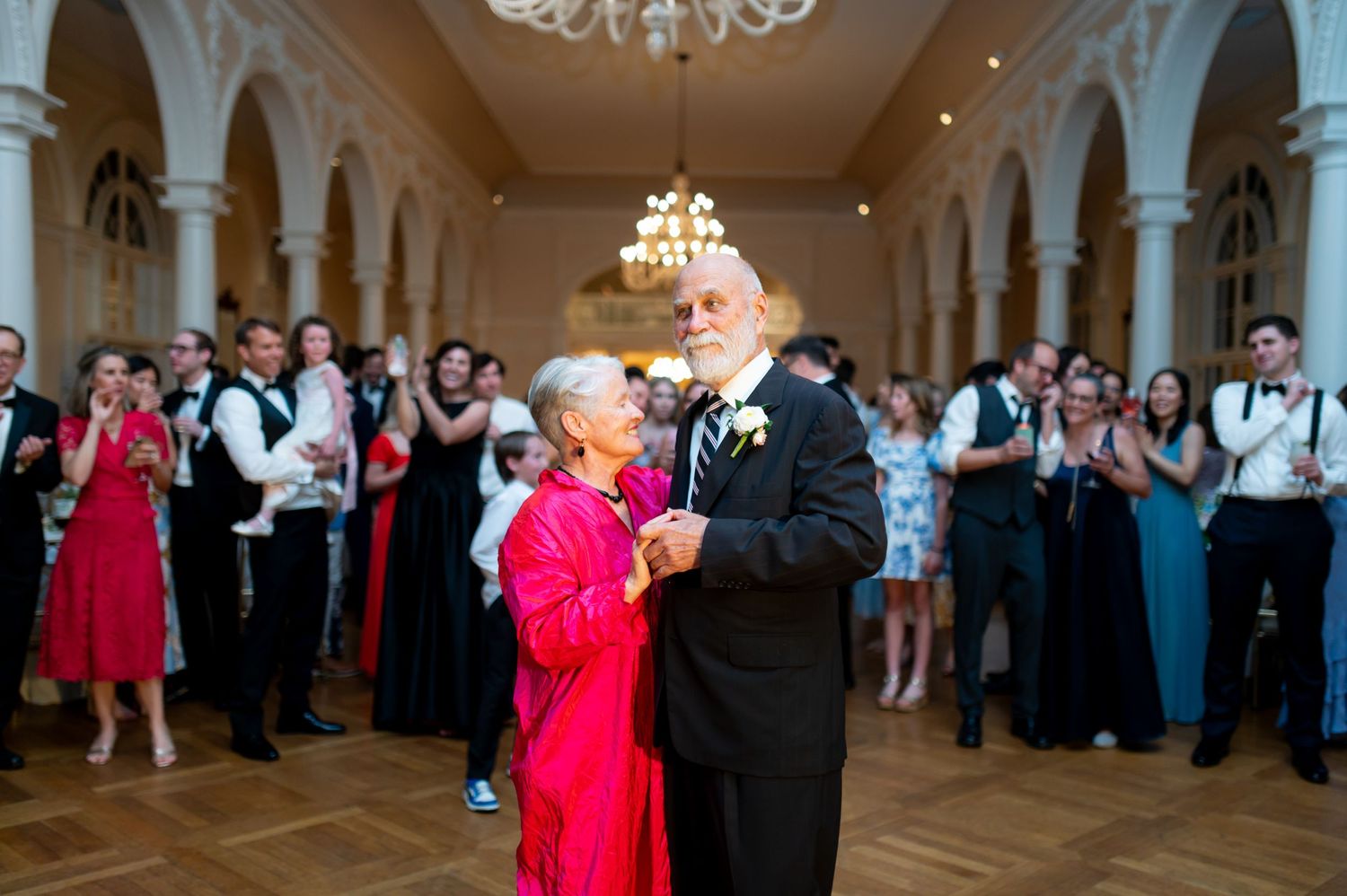 Elegant ballroom dance scene with chandeliers and arched columns during formal celebration.