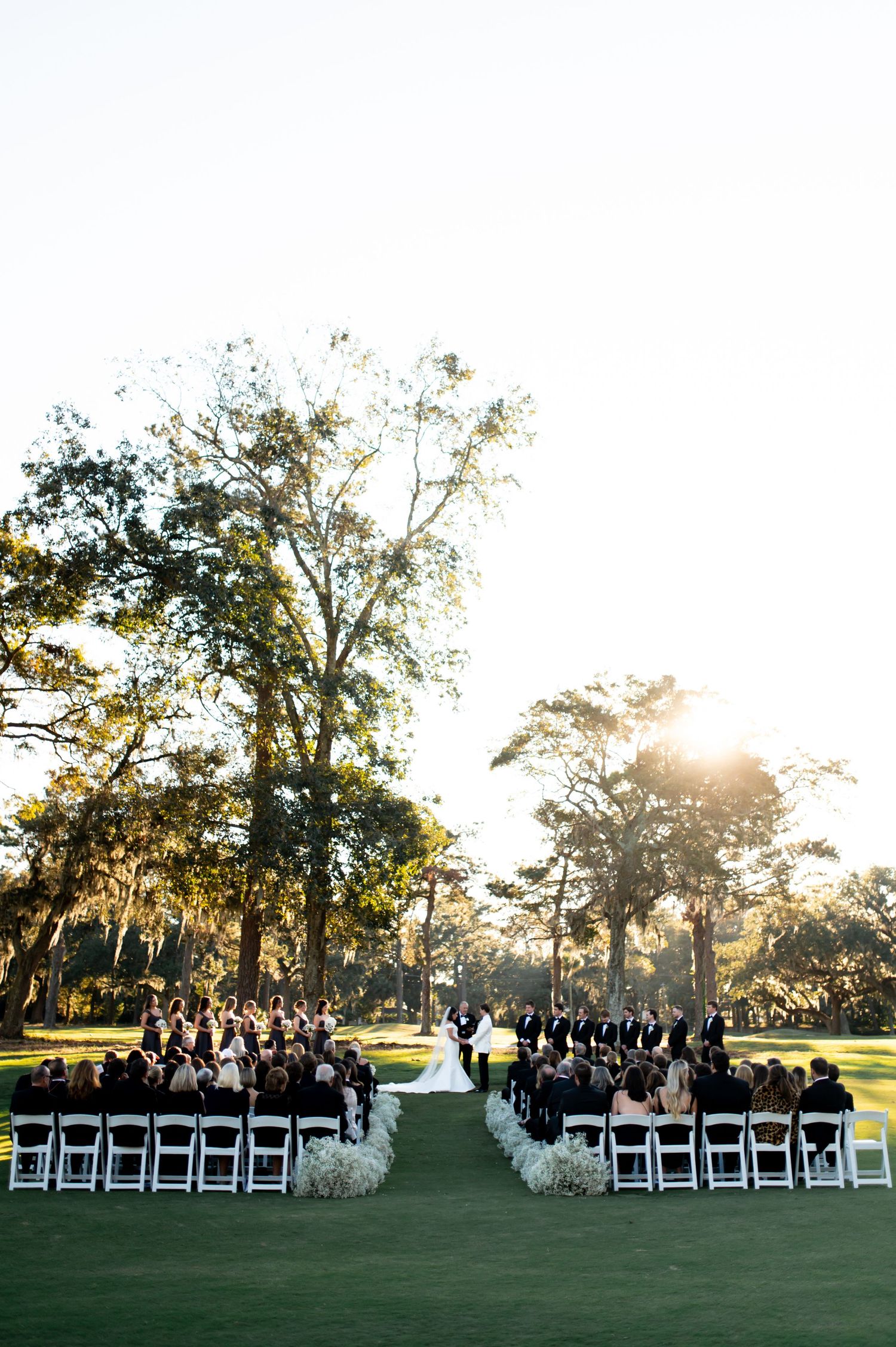 Sunset wedding ceremony setup with white chairs and floral arrangements under Spanish moss-draped trees.