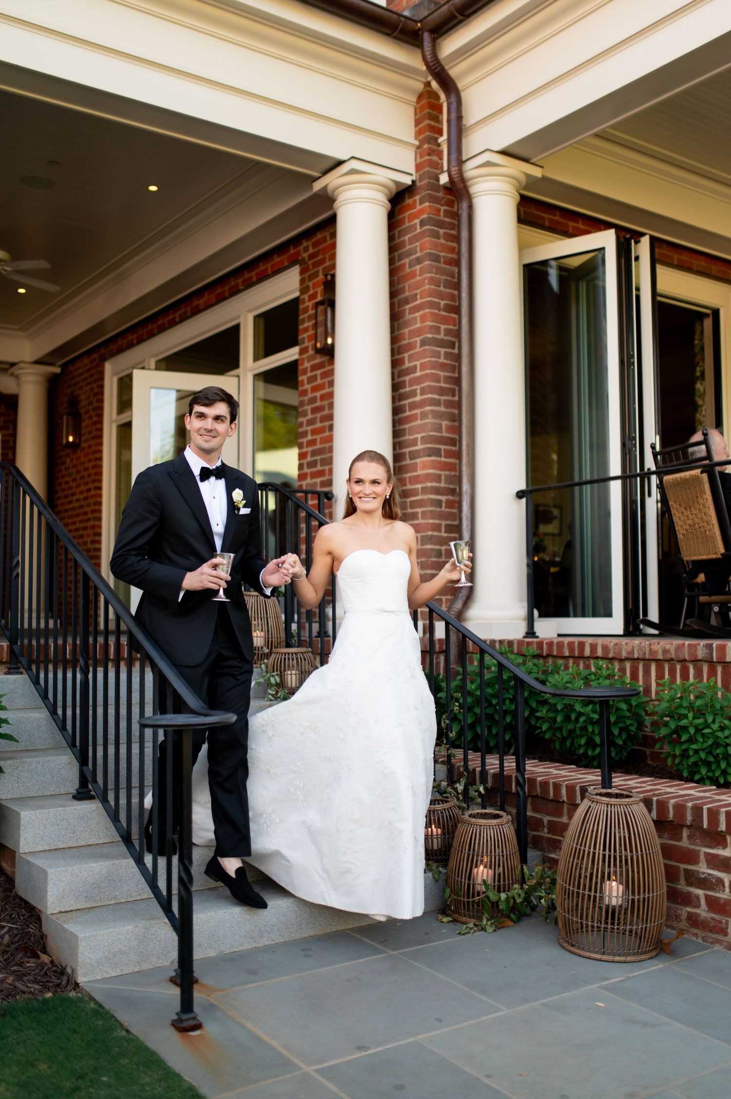 Bride and groom toast champagne on steps of elegant brick venue with white columns and lantern decor.