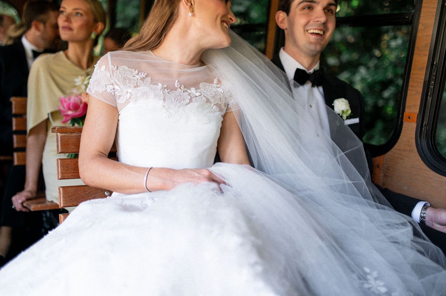 Close-up of bride in lace cap-sleeve gown and veil seated beside smiling groom in tuxedo.