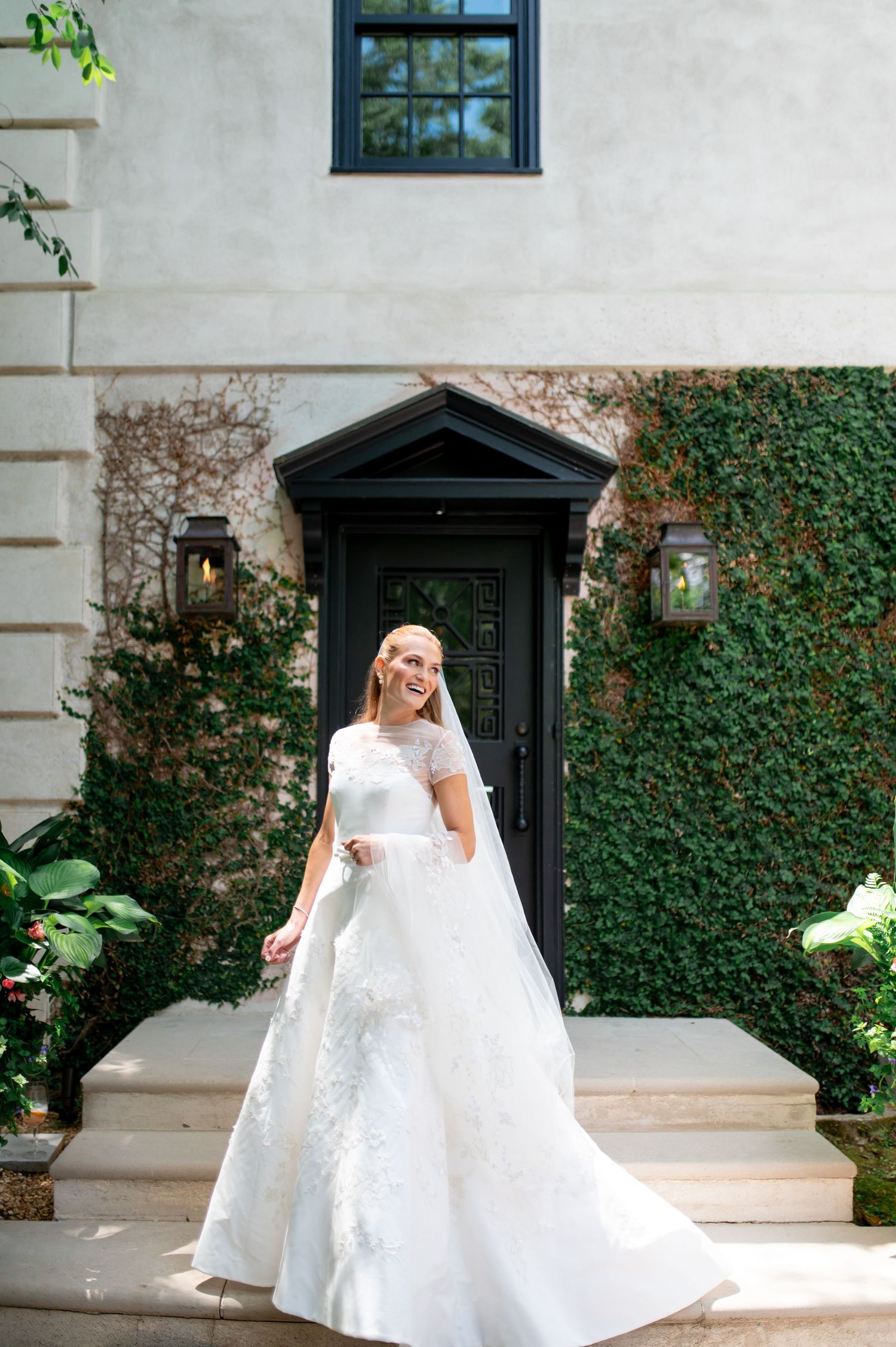 Smiling bride in white lace wedding gown with veil stands before ivy-covered wall and black door.