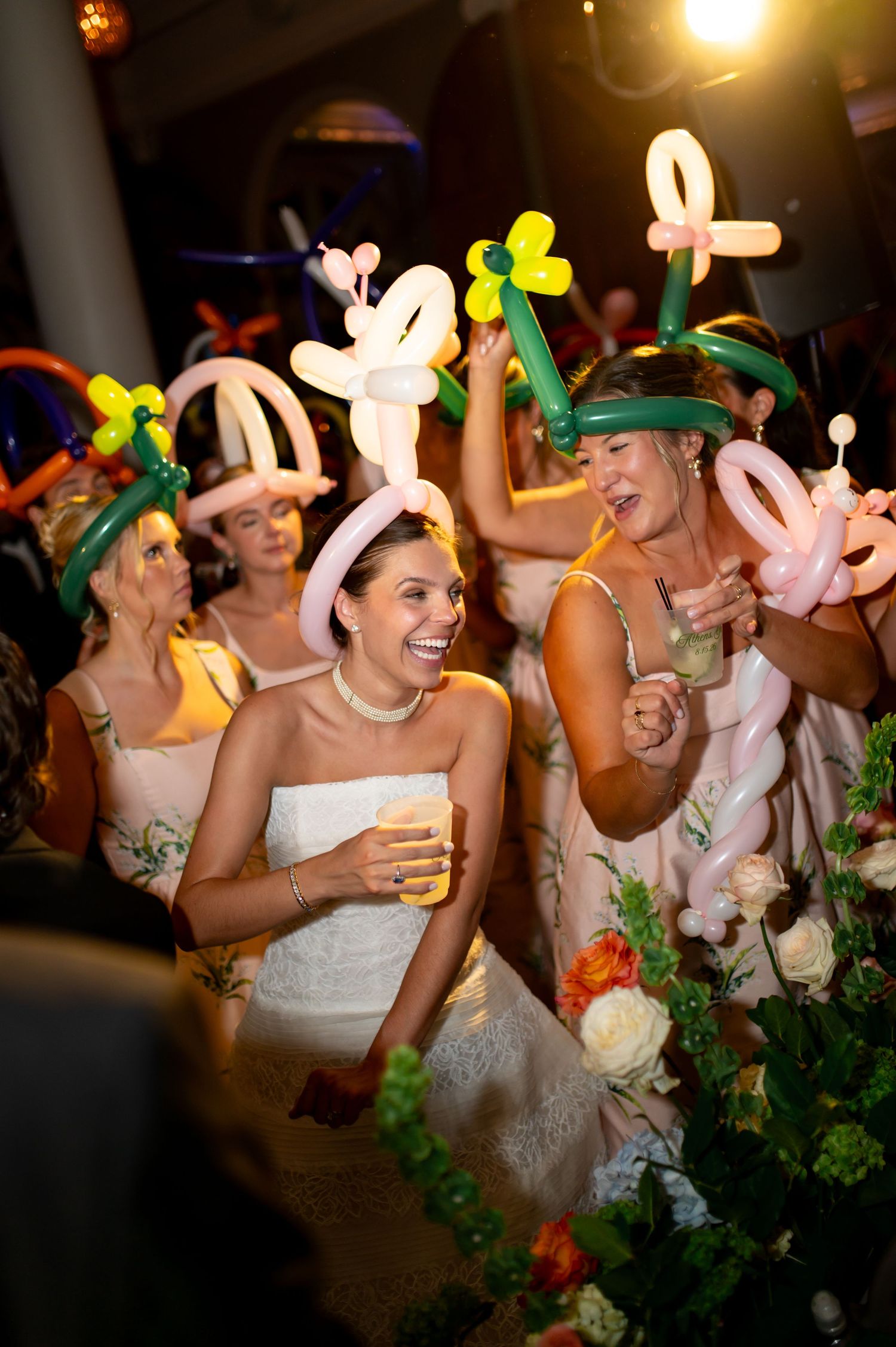 Bridesmaids wearing colorful balloon animal hats laugh and celebrate at a lively wedding reception.