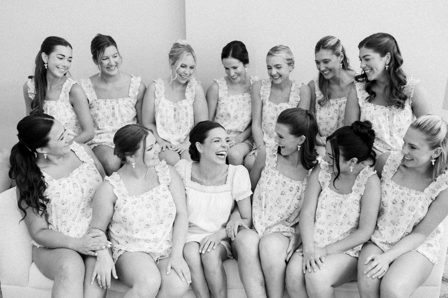 Black and white photo of a bride surrounded by smiling bridesmaids in matching floral rompers during wedding preparations.