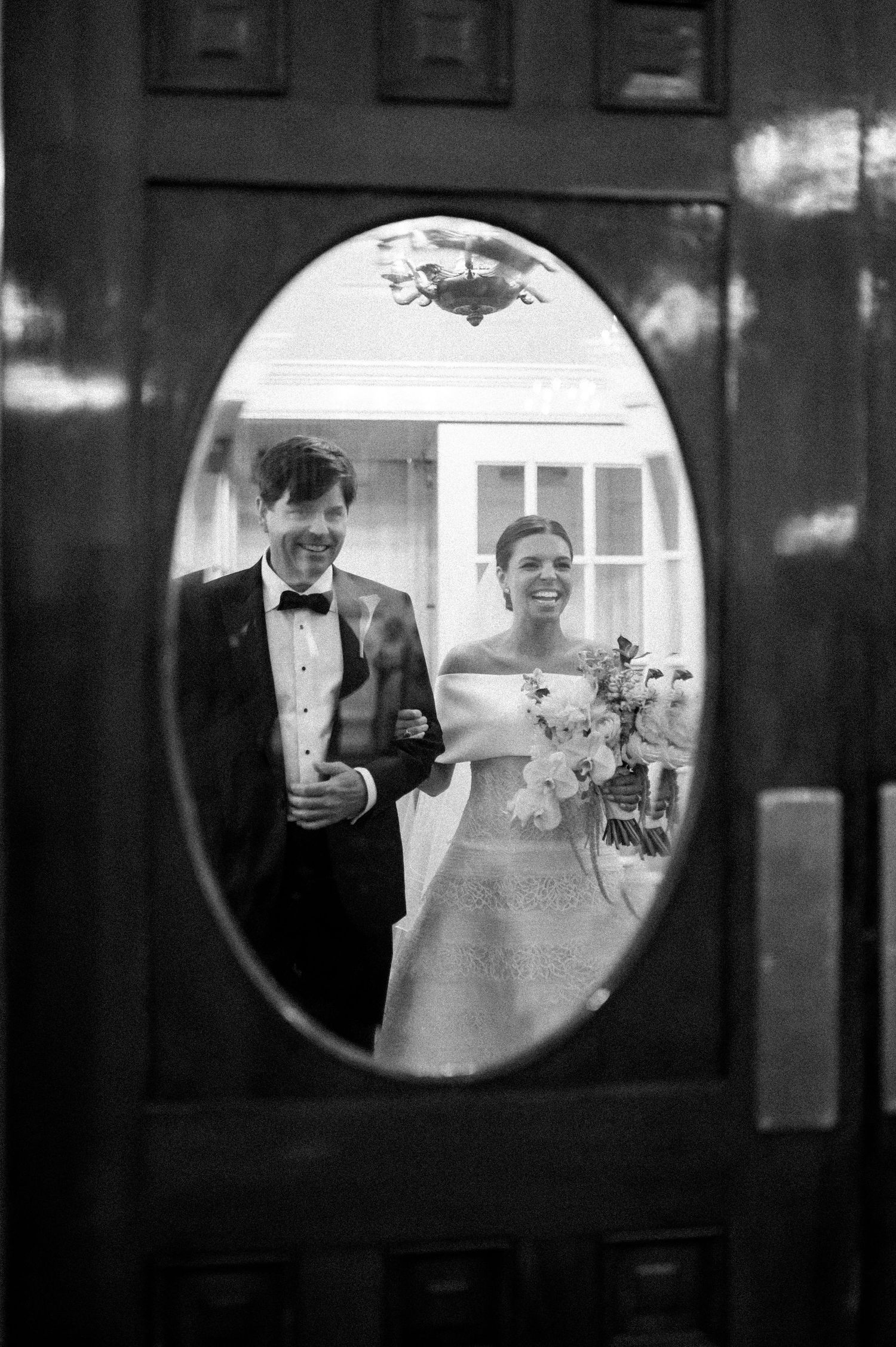 Bride and groom reflected in oval door mirror, bride holding bouquet in off-shoulder gown.