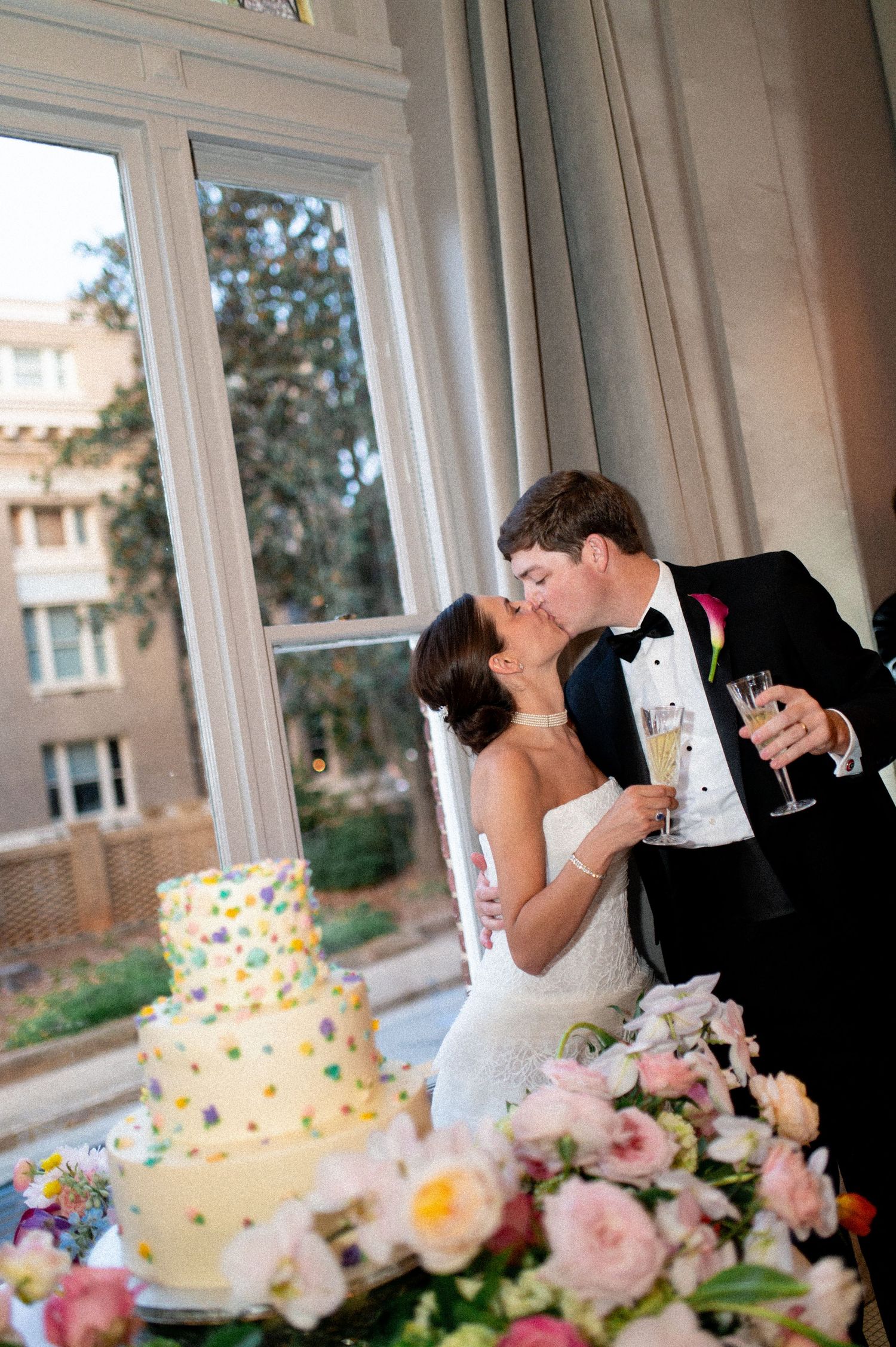 Bride and groom kiss beside a colorful polka dot wedding cake with champagne glasses at their reception.
