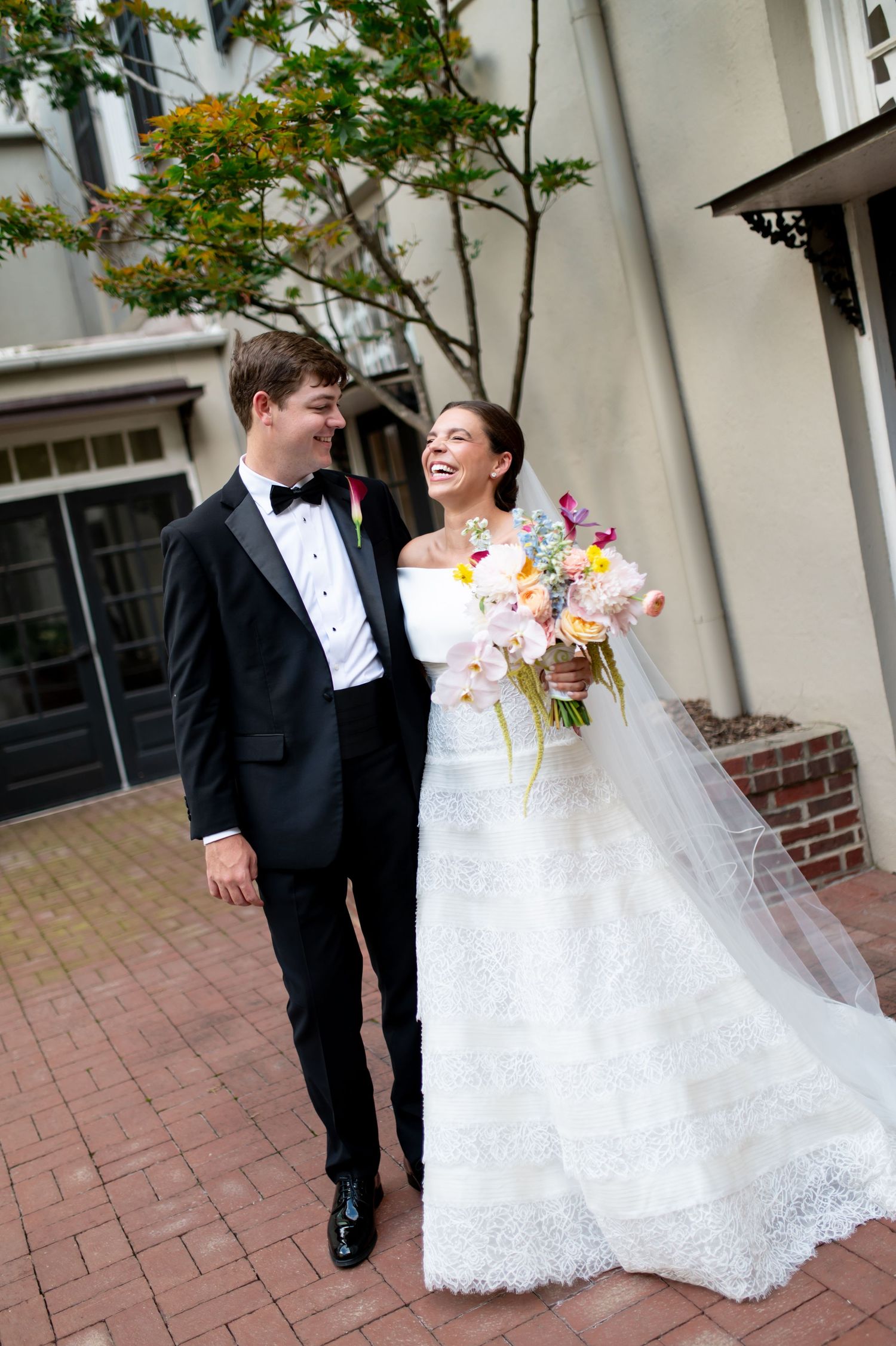 Bride in off-shoulder lace gown holding colorful bouquet walks with tuxedo-clad groom on brick courtyard.