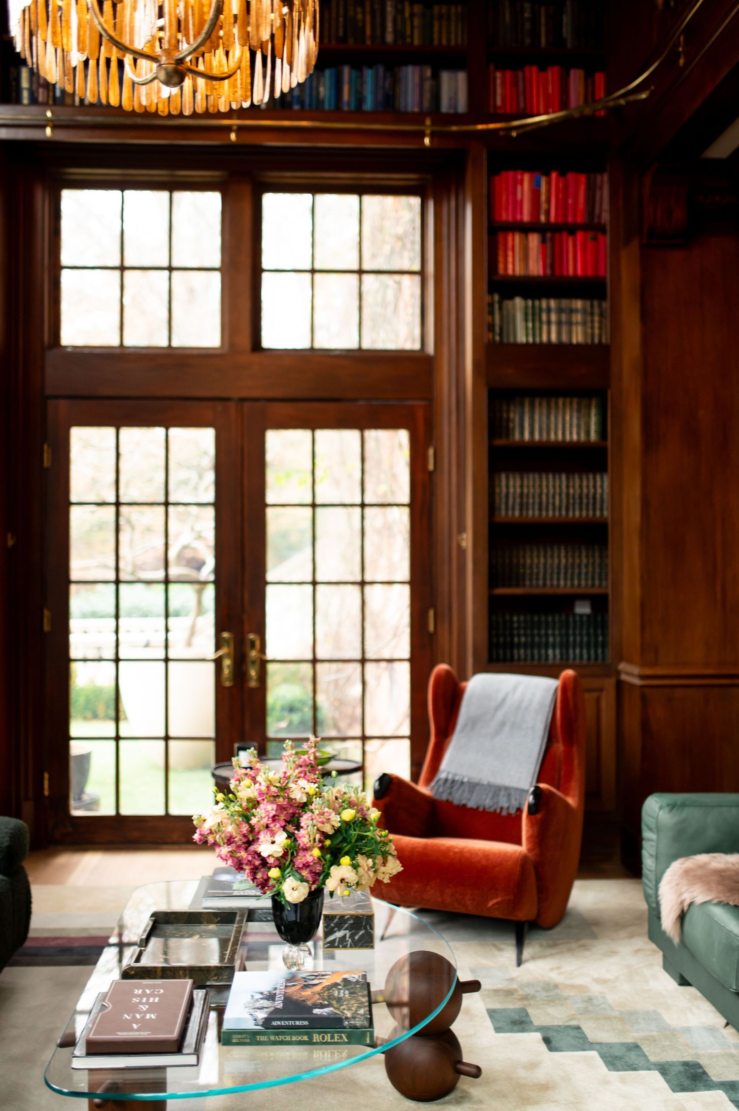 Cozy library with wood paneling, orange velvet armchair, glass coffee table, and built-in bookshelves by French doors.