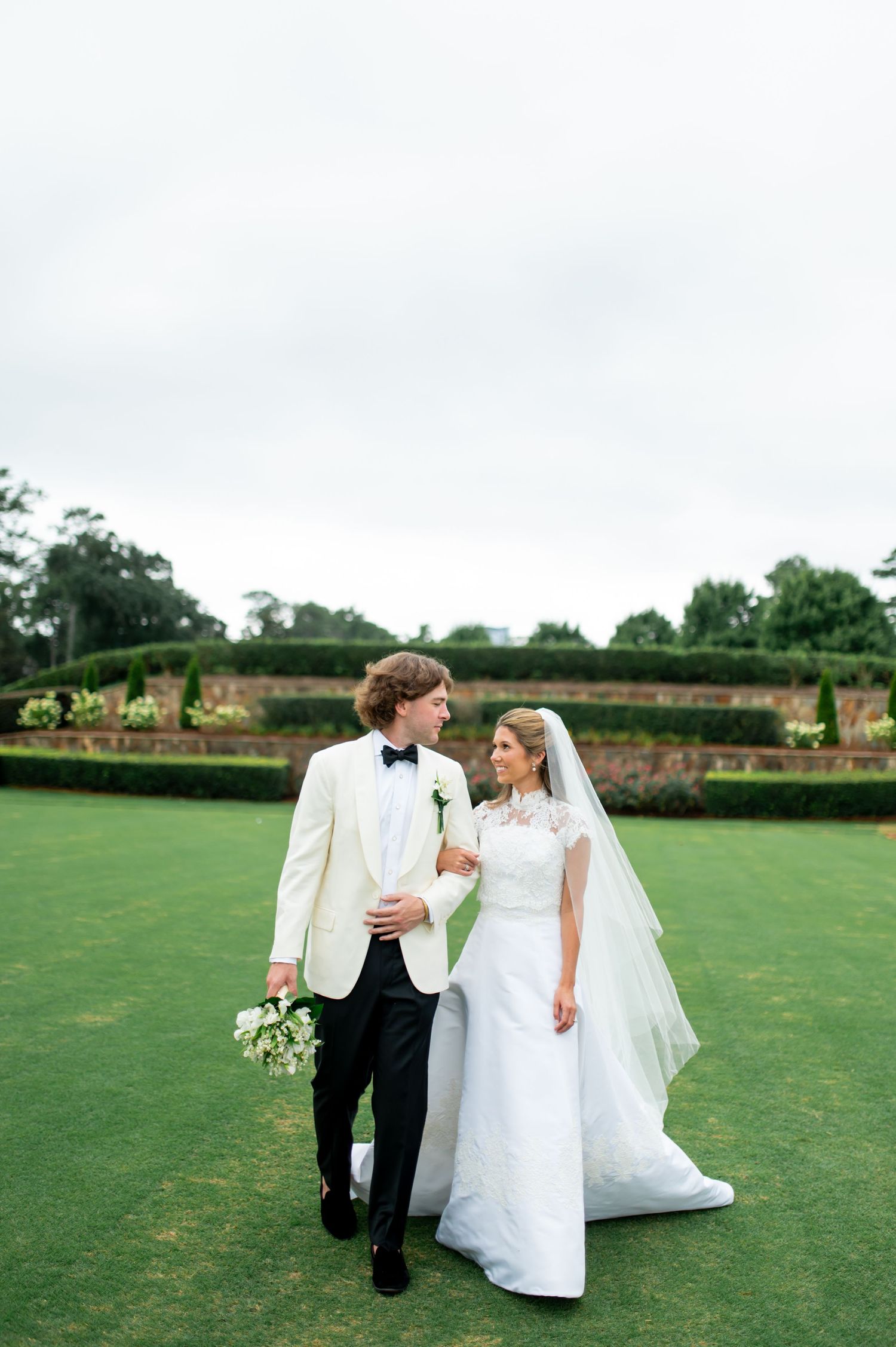 Newlyweds walk together on a manicured lawn in classic white wedding attire with a bouquet of white flowers.