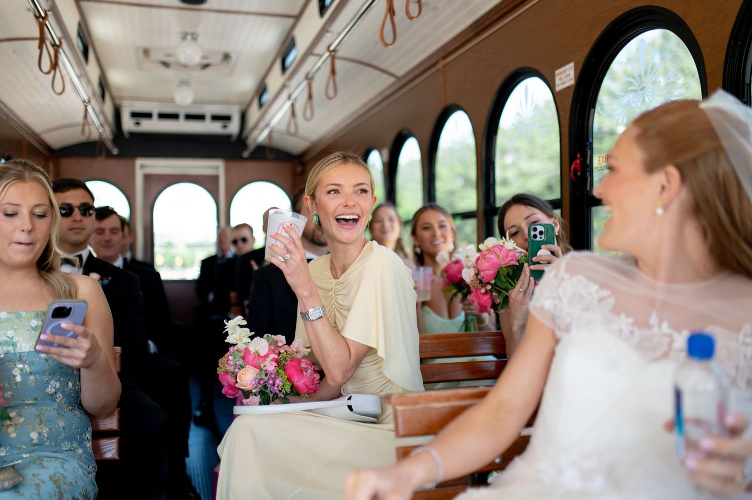 Joyful wedding guests celebrate on a vintage trolley with pink bouquets and phones.
