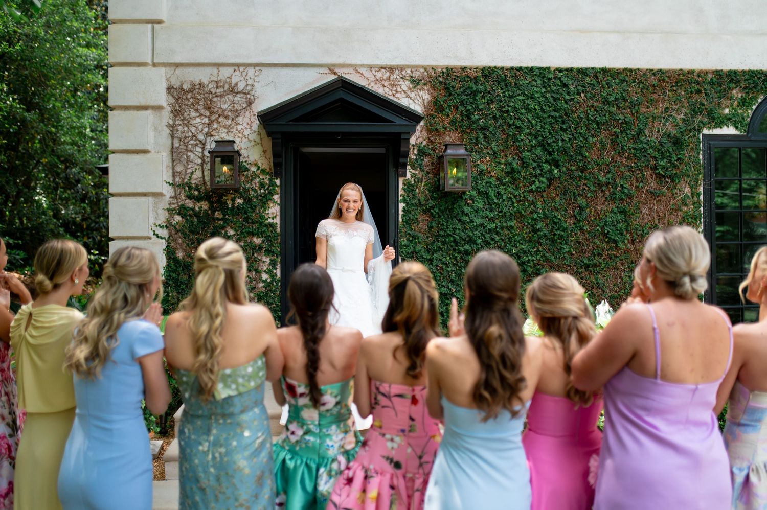 Bride in white gown and veil revealed to cheering bridesmaids in colorful dresses outside ivy-covered venue entrance.