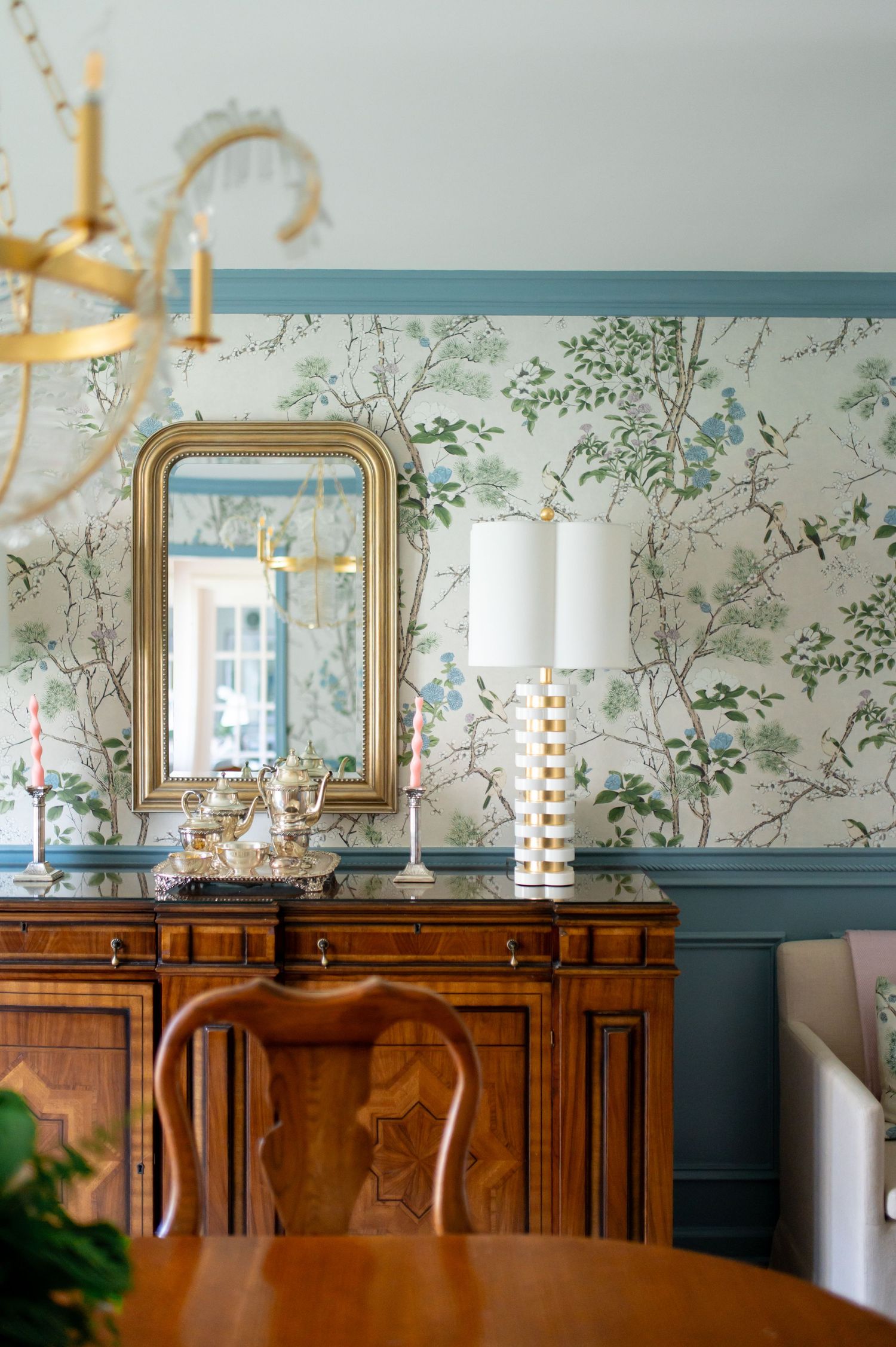 Elegant dining room with floral chinoiserie wallpaper, antique wood sideboard, gold mirror, and brass chandelier.