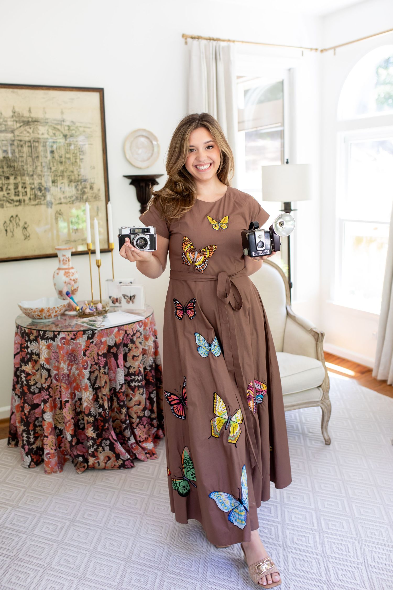 Woman in brown butterfly maxi dress holding two vintage cameras in elegant living room.