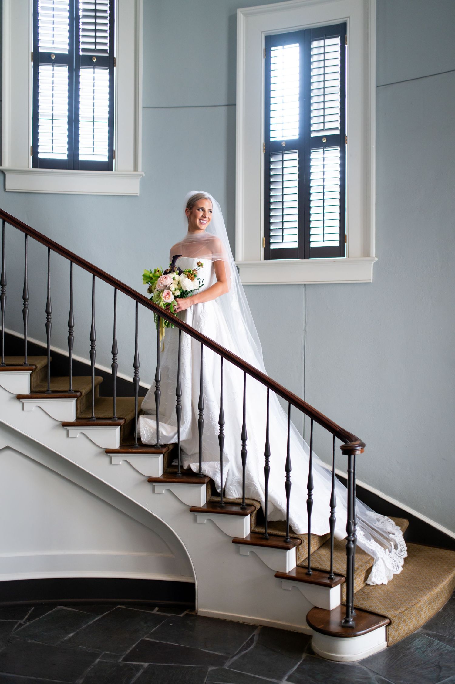 A stunning wedding dress photographed on an elegant staircase with white walls and dark wood railings.