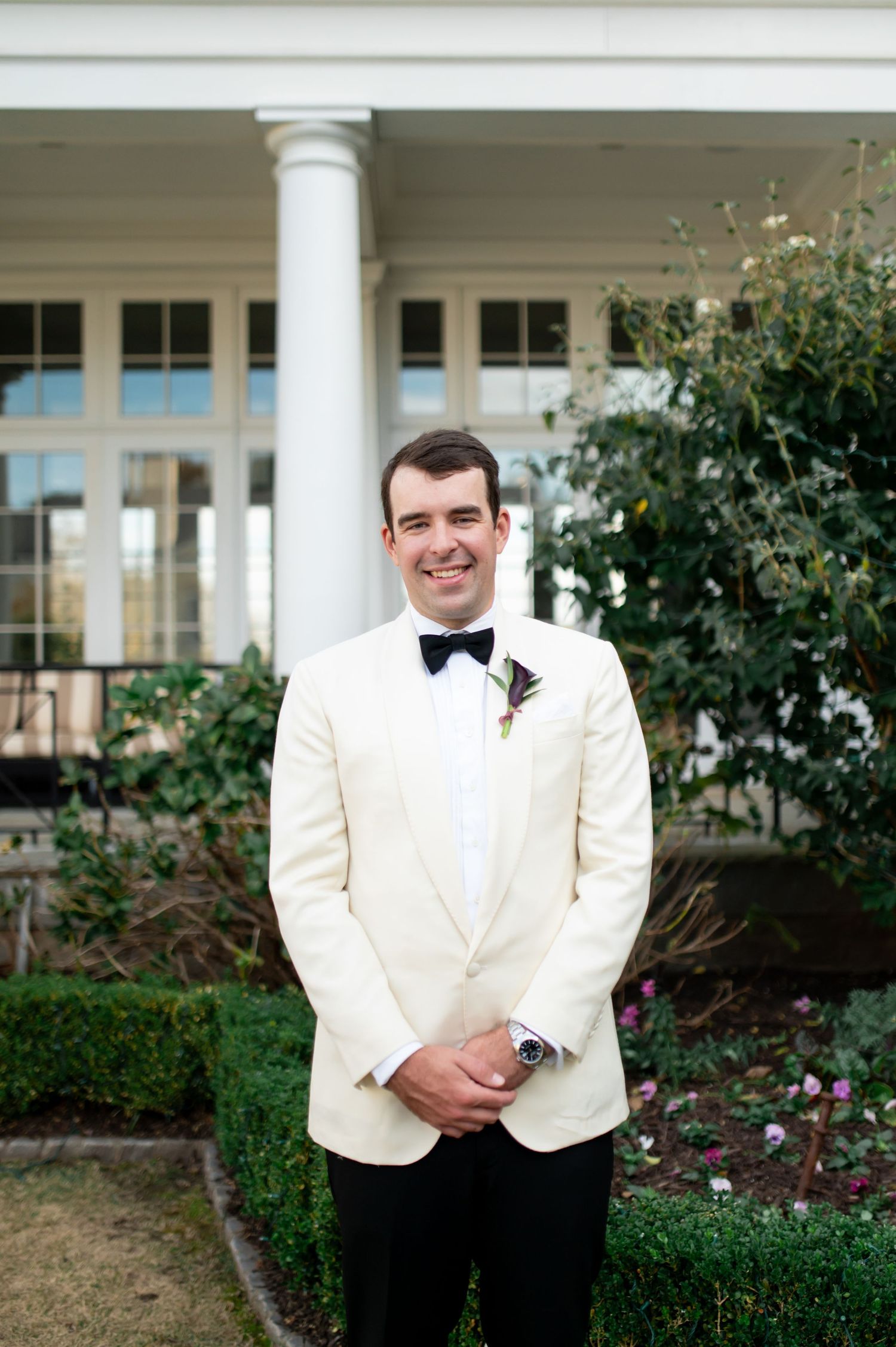 Groom in white dinner jacket and black bow tie poses in front of columned building.