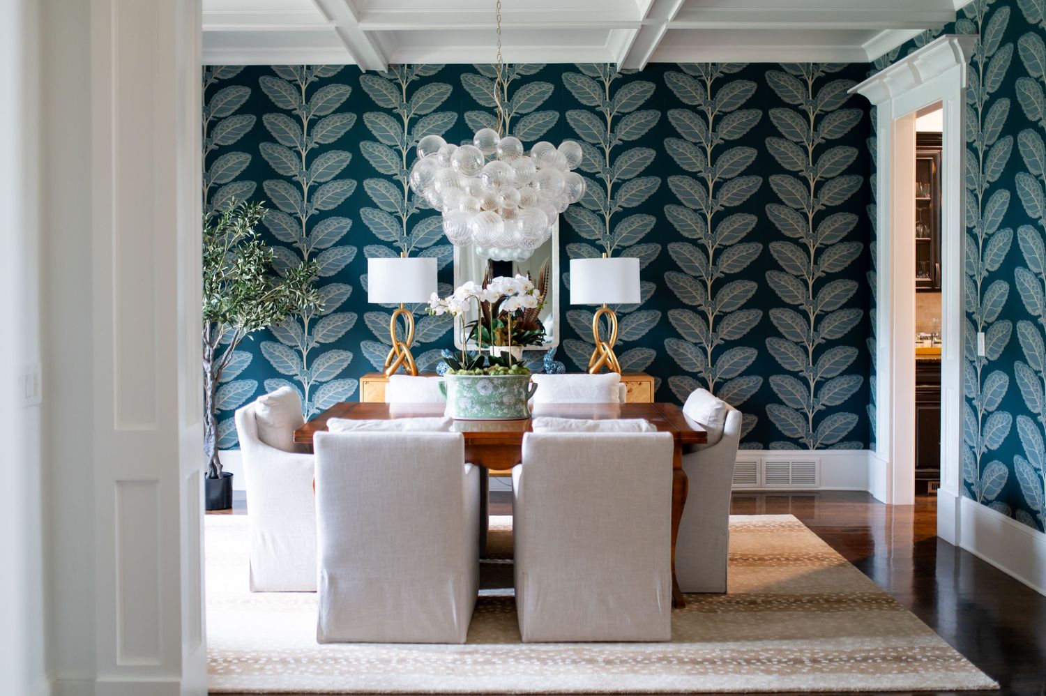 Modern dining room features bold teal leaf wallpaper, white upholstered chairs, and an elegant crystal chandelier.